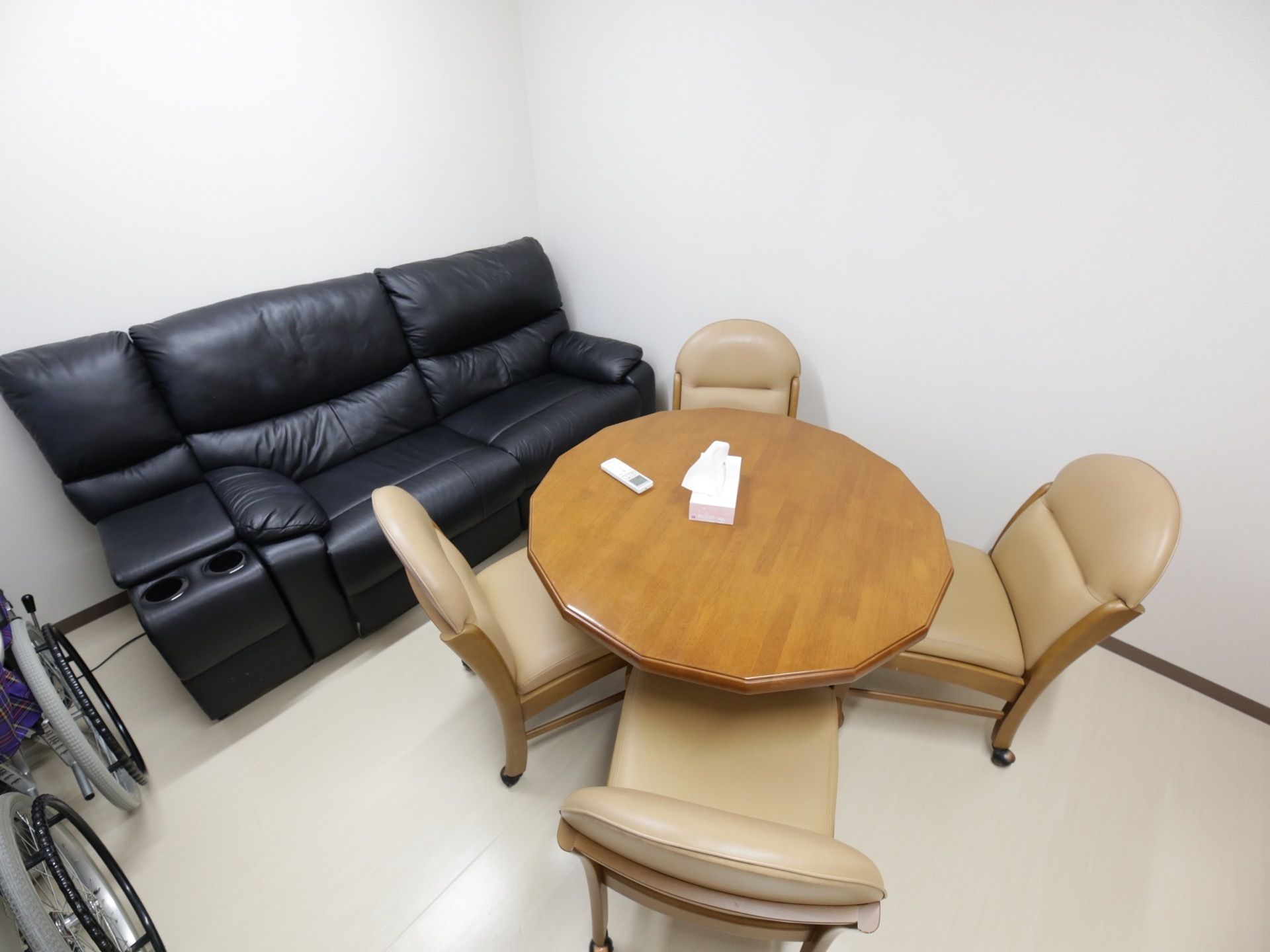 木製の丸いテーブルの周りに、4脚のベージュの椅子が並べられている。側の壁際には、2人掛けの黒いソファが置かれている。