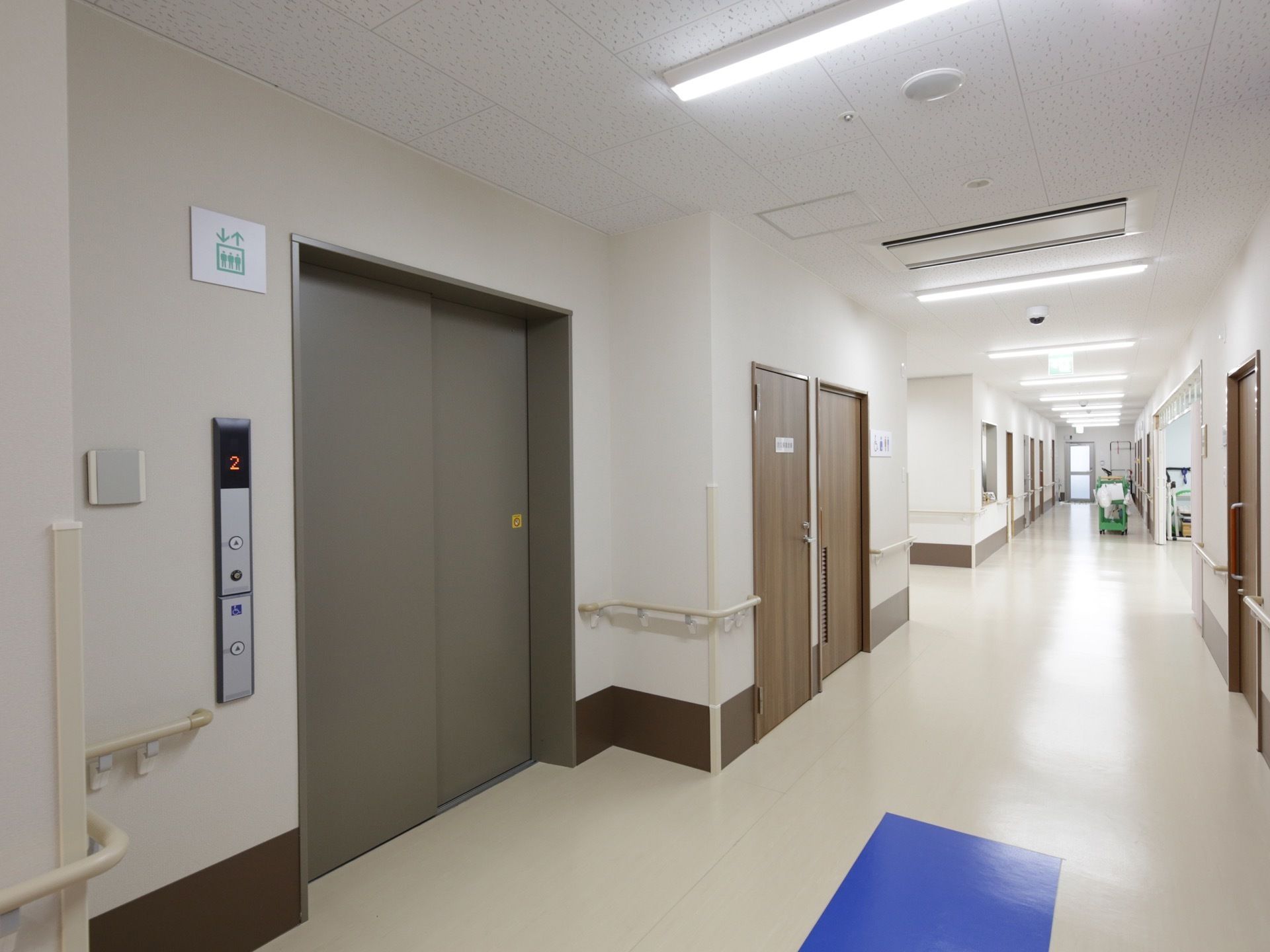 ライトグレーの床と白い壁の廊下の端にエレベーターがある。両側には居室のドアが並び、壁には手すりがある。