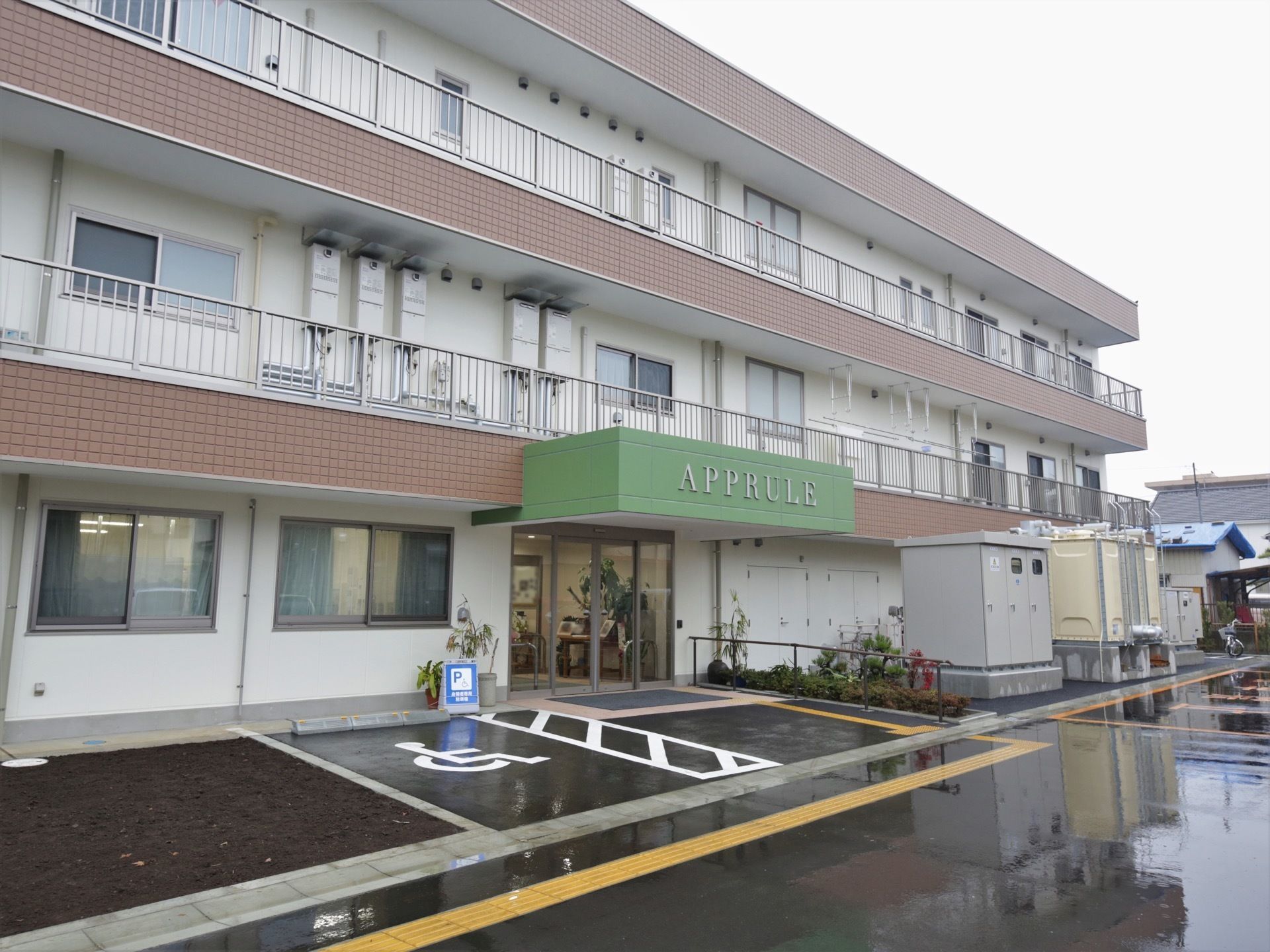 3階建ての大きな建物がある。前面部分には窓があり、柵の付いたベランダになっている。地面は舗装され、駐車場がある。