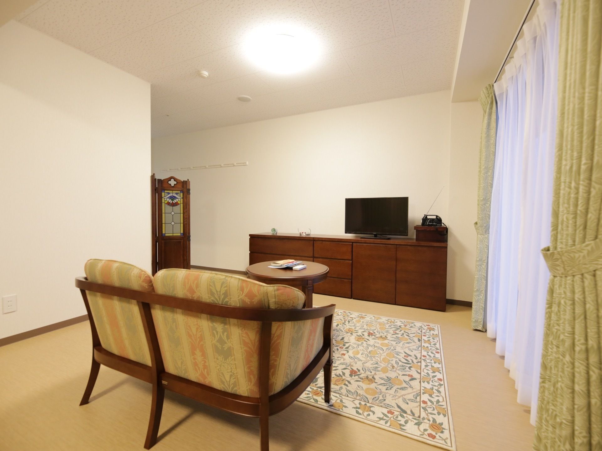 白い壁とベージュの床の居室がある。床には絨毯が敷かれ、レースのカーテンの掛かった窓がある。ソファ、テーブル、テレビの乗った棚がある。