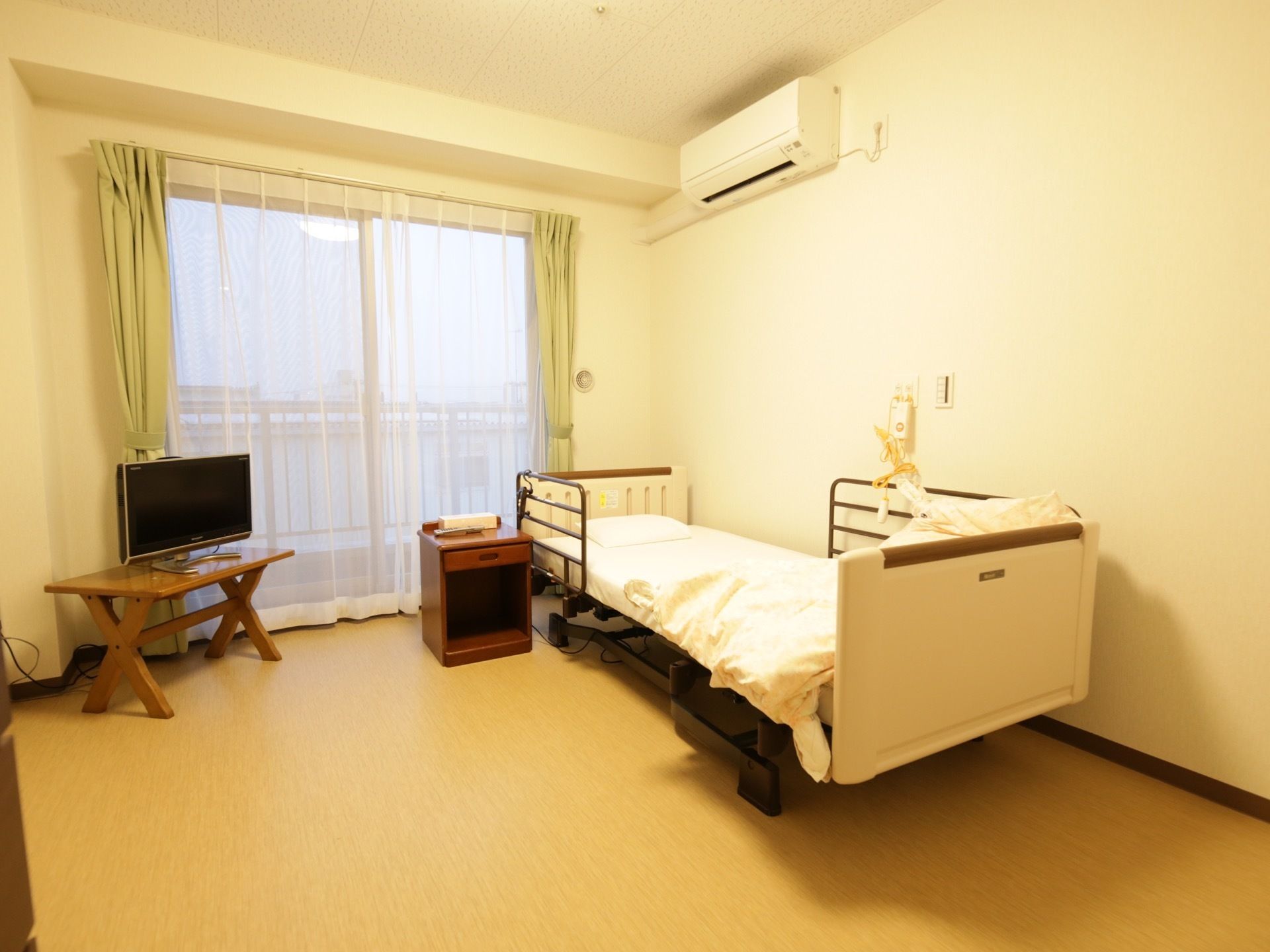 白い壁とベージュの床の居室である。カーテンの掛かった窓があり、壁にはエアコンが設置されている。柵の付いた介護用ベッドがある。
