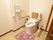 白い壁とベージュの床のトイレがある。便座にはひじ掛けがあり、壁には手すりがある。温水洗浄便座が付いている。
