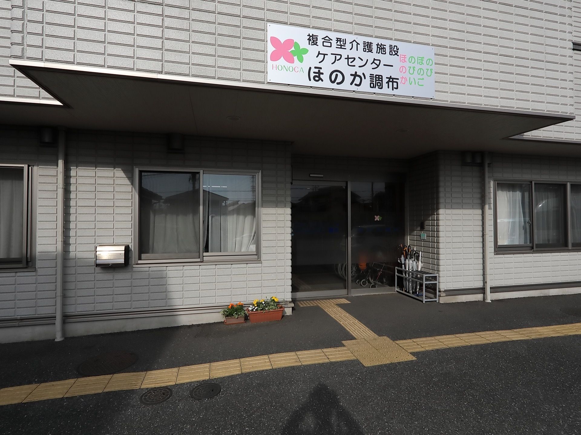 ガラス張りの自動ドアの玄関である。上部には屋根を設け、その上に施設の看板を掲げている。地面には、点字ブロックを設置している。