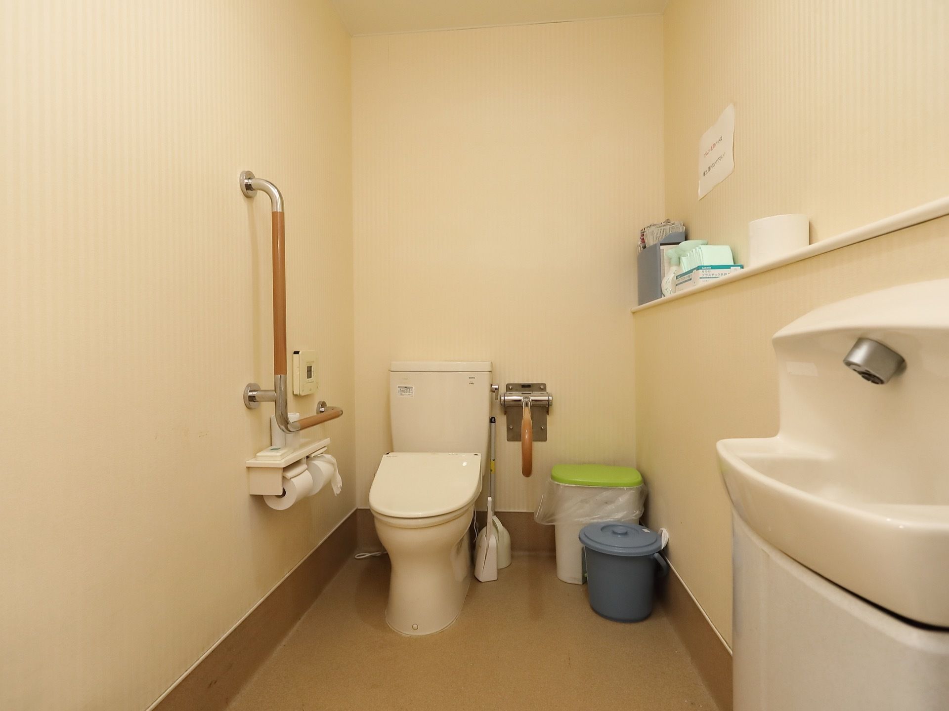 白を基調とし、床にブラウンを取り入れたトイレである。便座の両側に手すりを備え、前方の壁際に洗面台を設置している。