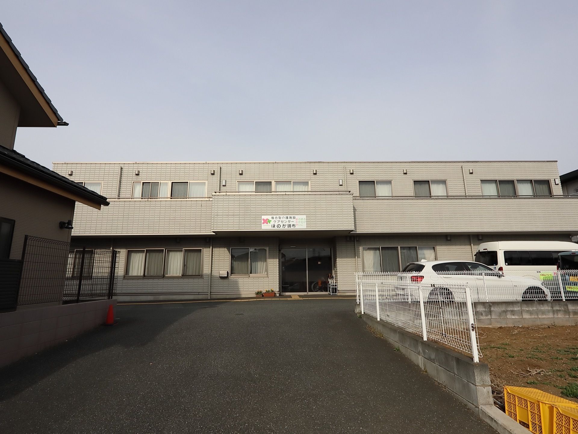 アスファルトの道路を真っ直ぐ進んだ所に佇む、２階建ての建物である。玄関の上部には、施設の看板を掲げている。横側に数台の車が停まっている。
