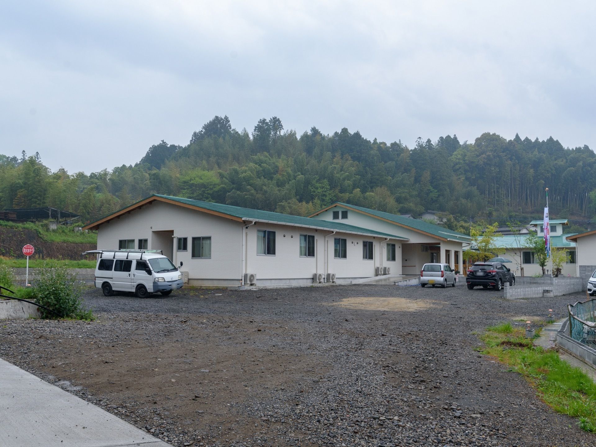 平屋建ての施設の前には広い駐車スペースがあり、数台の車が置いてある。後方には、自然豊かな山並みが見える。