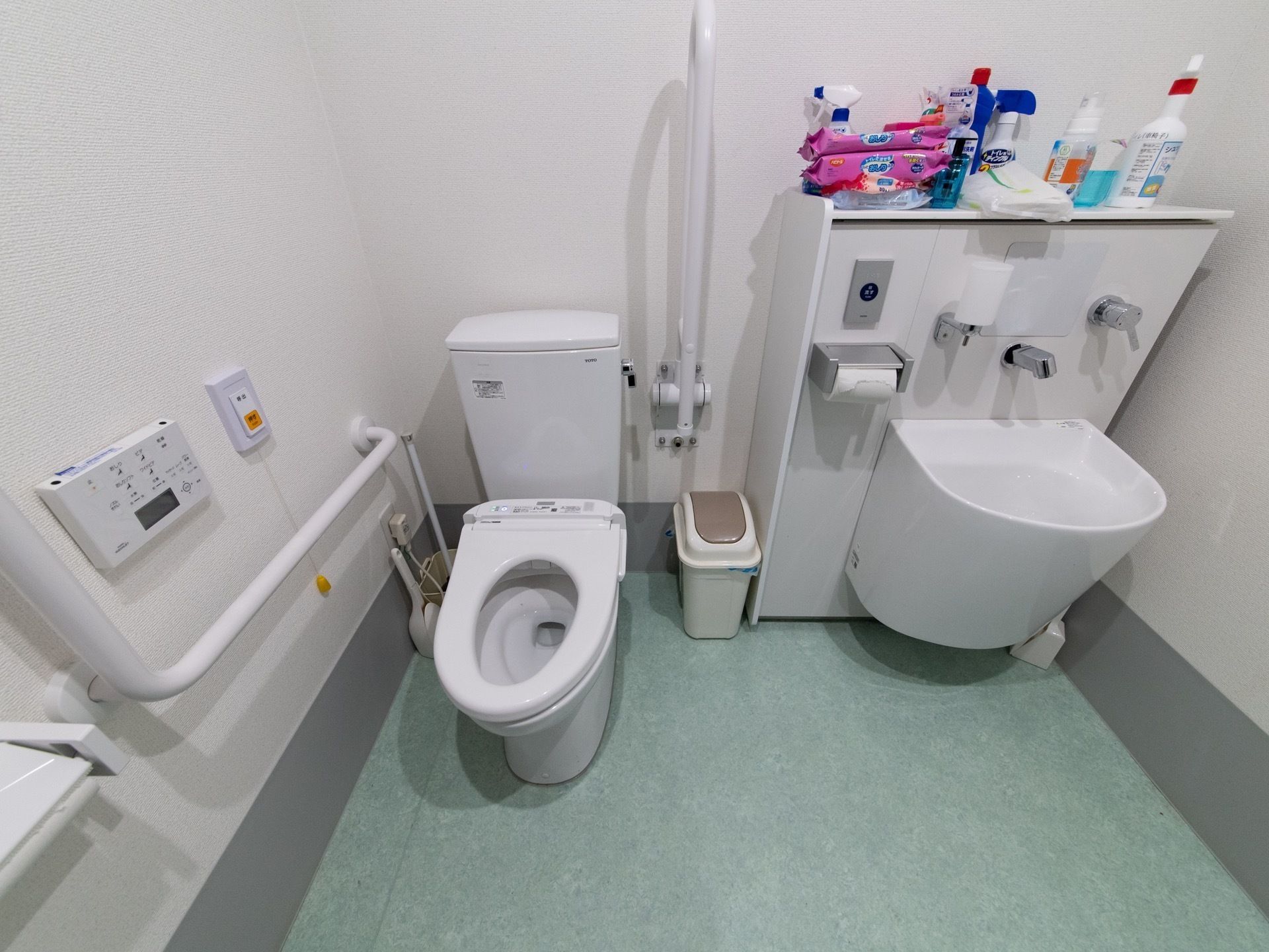 広々としたトイレスペースである。便座の傍に可動式の手すりを備え、その横に大きめの洗面台を設けている。壁にはエル型の手すりを設置している。