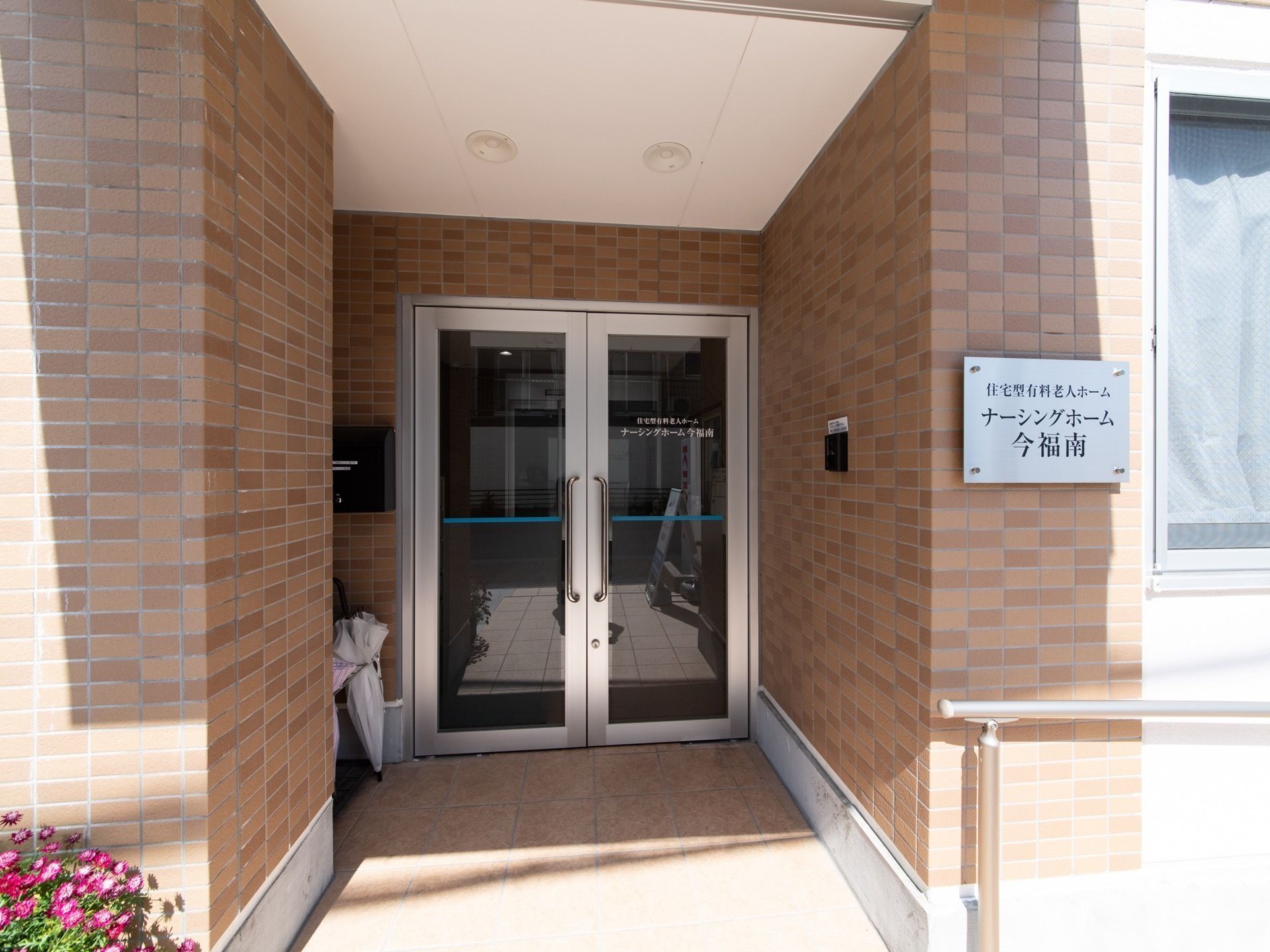 レンガ調の壁に囲まれたガラス張りの玄関ドアがある。前方横の外壁にシルバーのプレートに施設種類と施設名が書かれた表札を掲げている。