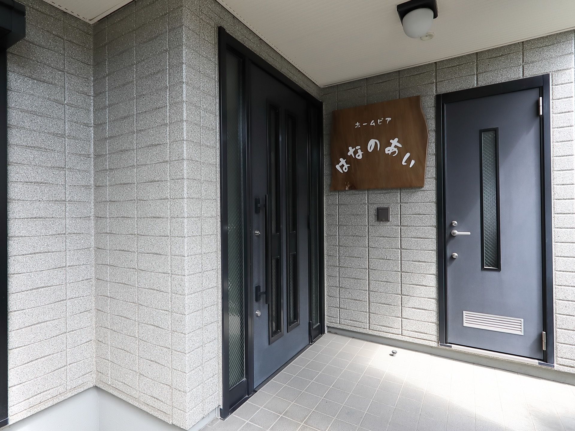 玄関は、大きな取っ手がついた開き戸になっている。勝手口との間の壁に、施設名称が書かれた看板が設置されている。
