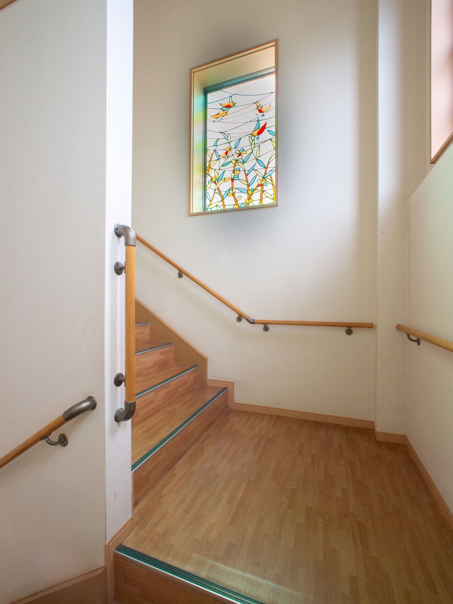 木目調の床の階段には、踊り場が設けられている。白い壁の両側には、木製の手すりが設置されている。小窓はステンドグラス窓になっている。