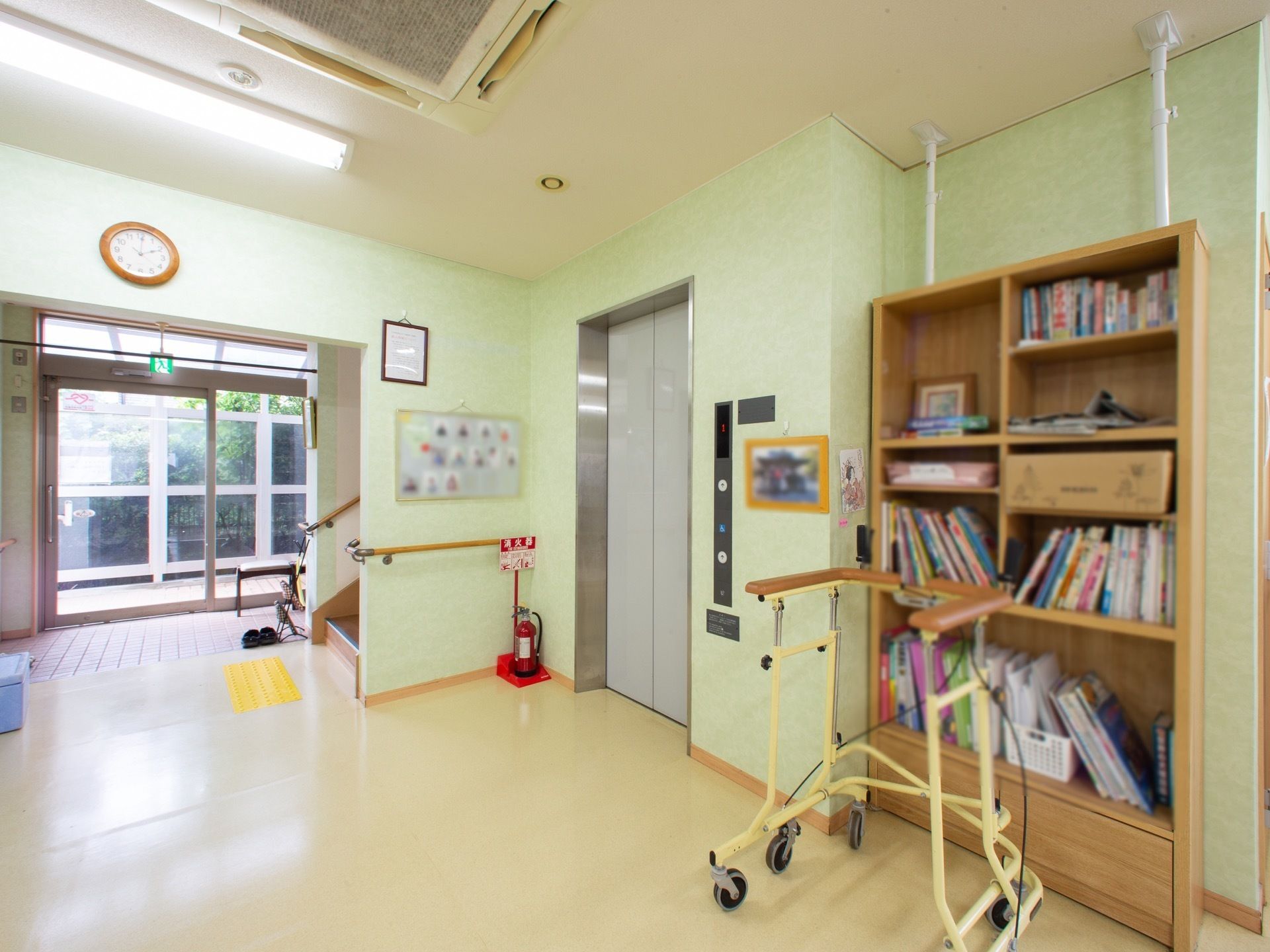 玄関を入ると、階段とエレベーターが併設されている。エレベータ横には本棚が設置されており、本棚の前には車輪付きの歩行器が置かれている。