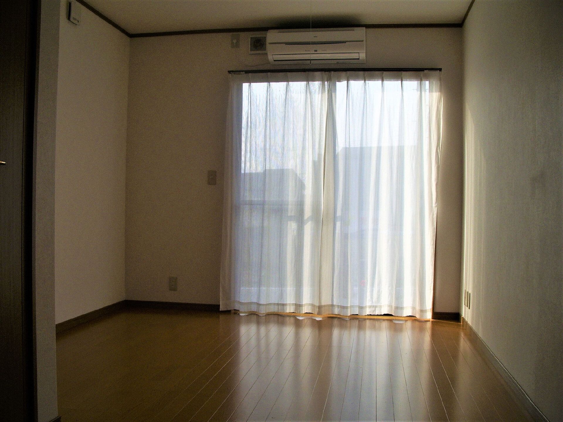 エアコンが付いたフローリングの部屋にはレースカーテンが付いた大きな窓があり、明るい雰囲気。