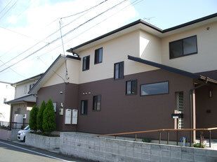 ベージュとブラウンの2階建ての建物は一般の住宅を大きくしたような外観。植物が植えられ、車も止められている。