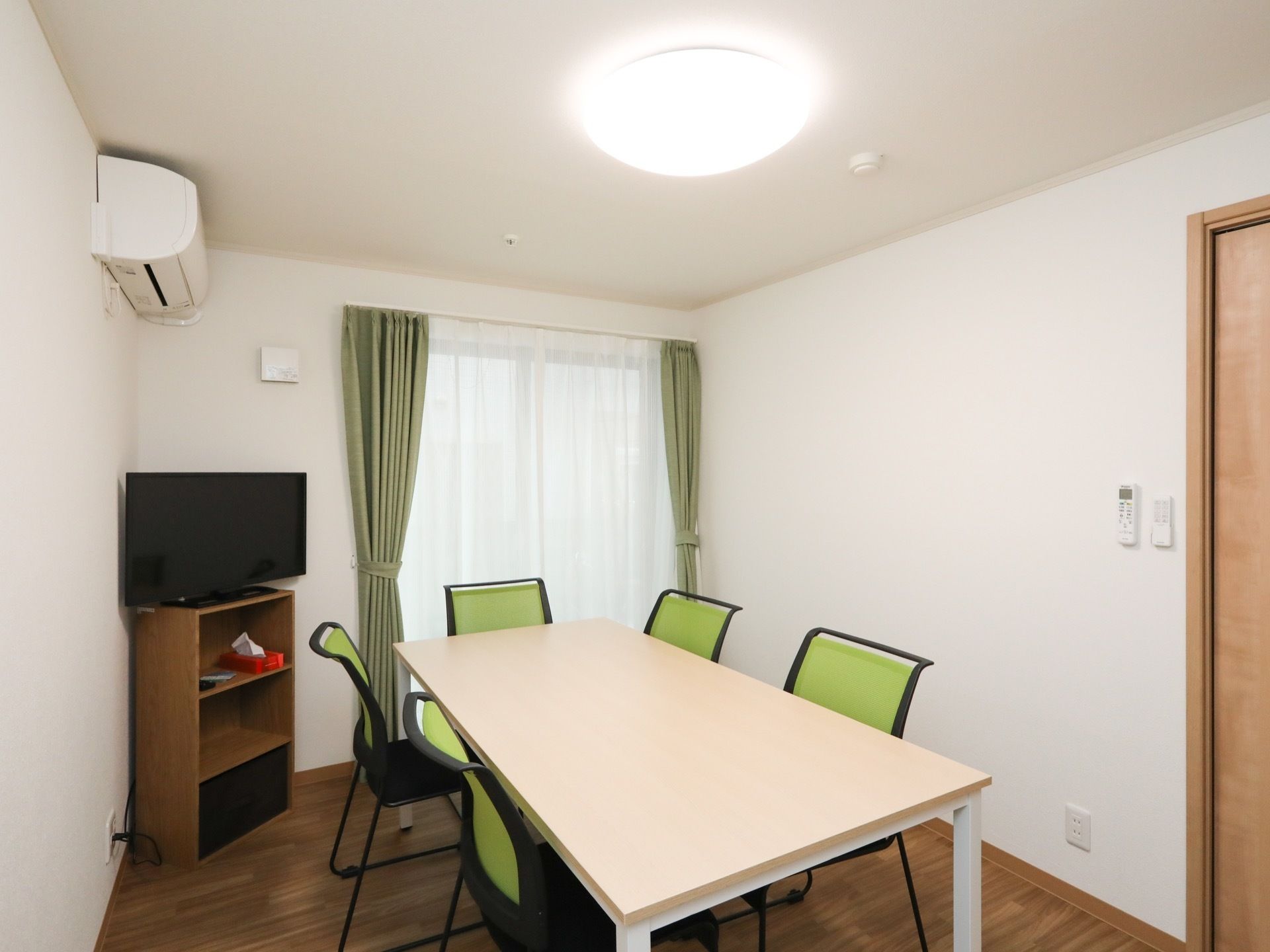 フローリングと白い壁の小部屋がある。カーテンの掛かった窓があり、中央にはテーブルと椅子が5脚置かれている。壁にはエアコンが設置されている。