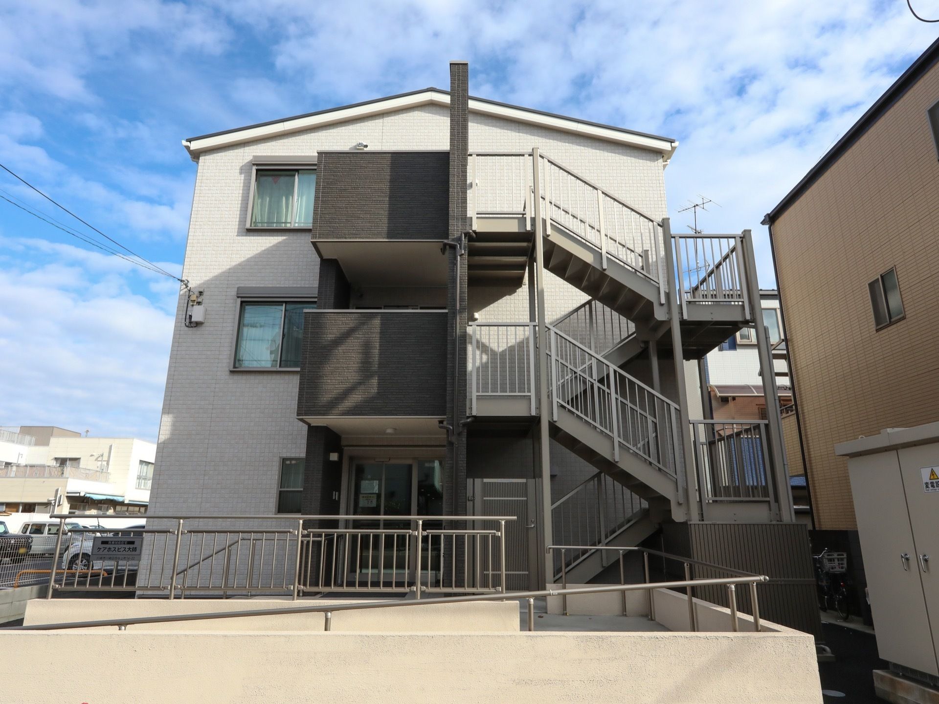 3階建ての建物がある。側面には外付けの非常階段が設置されている。玄関に向かってスロープと手すりが続いている。