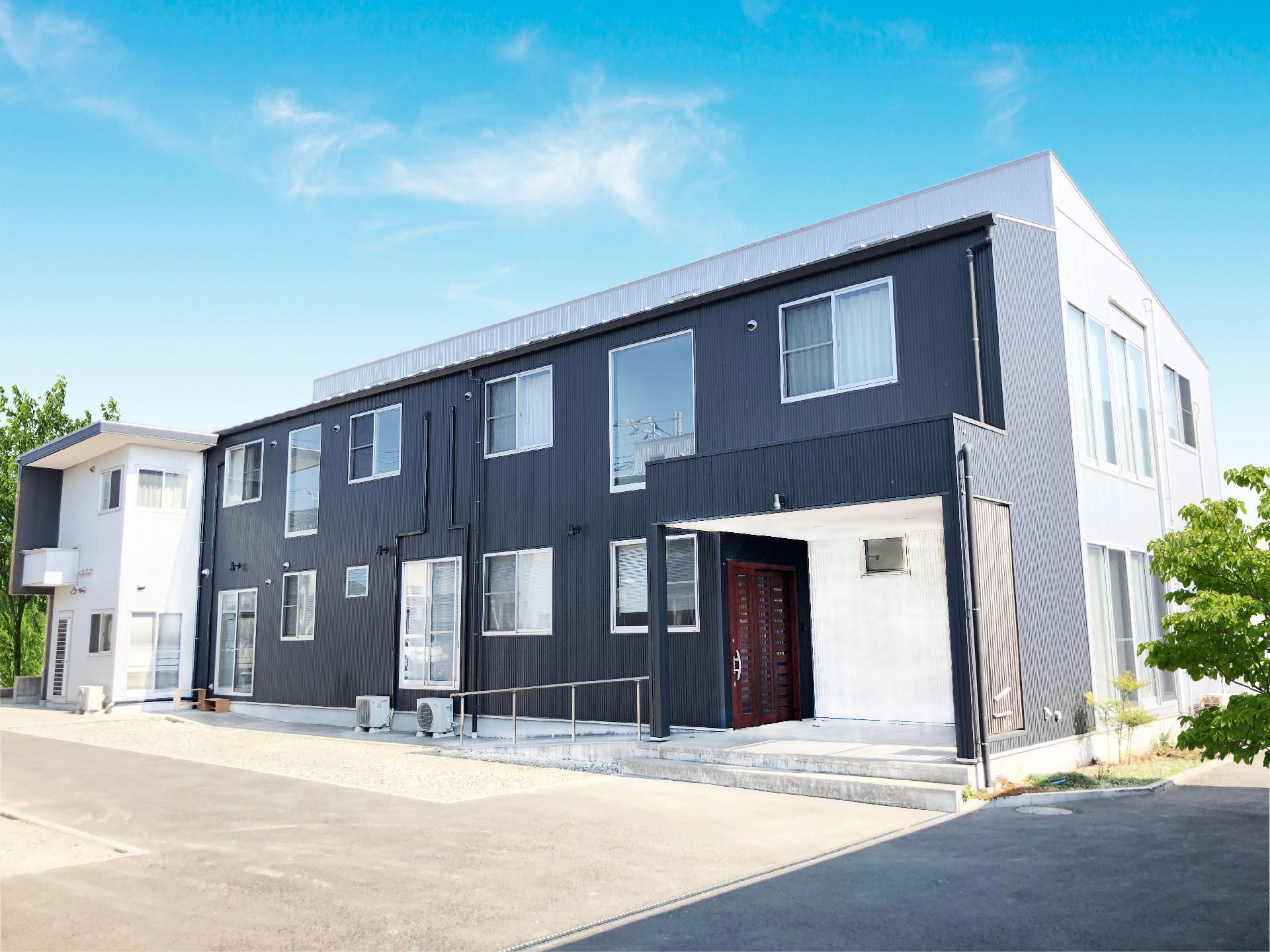緑に囲まれた環境。２階建ての建物は白と紺で玄関前には手すり付きのスロープがある。。