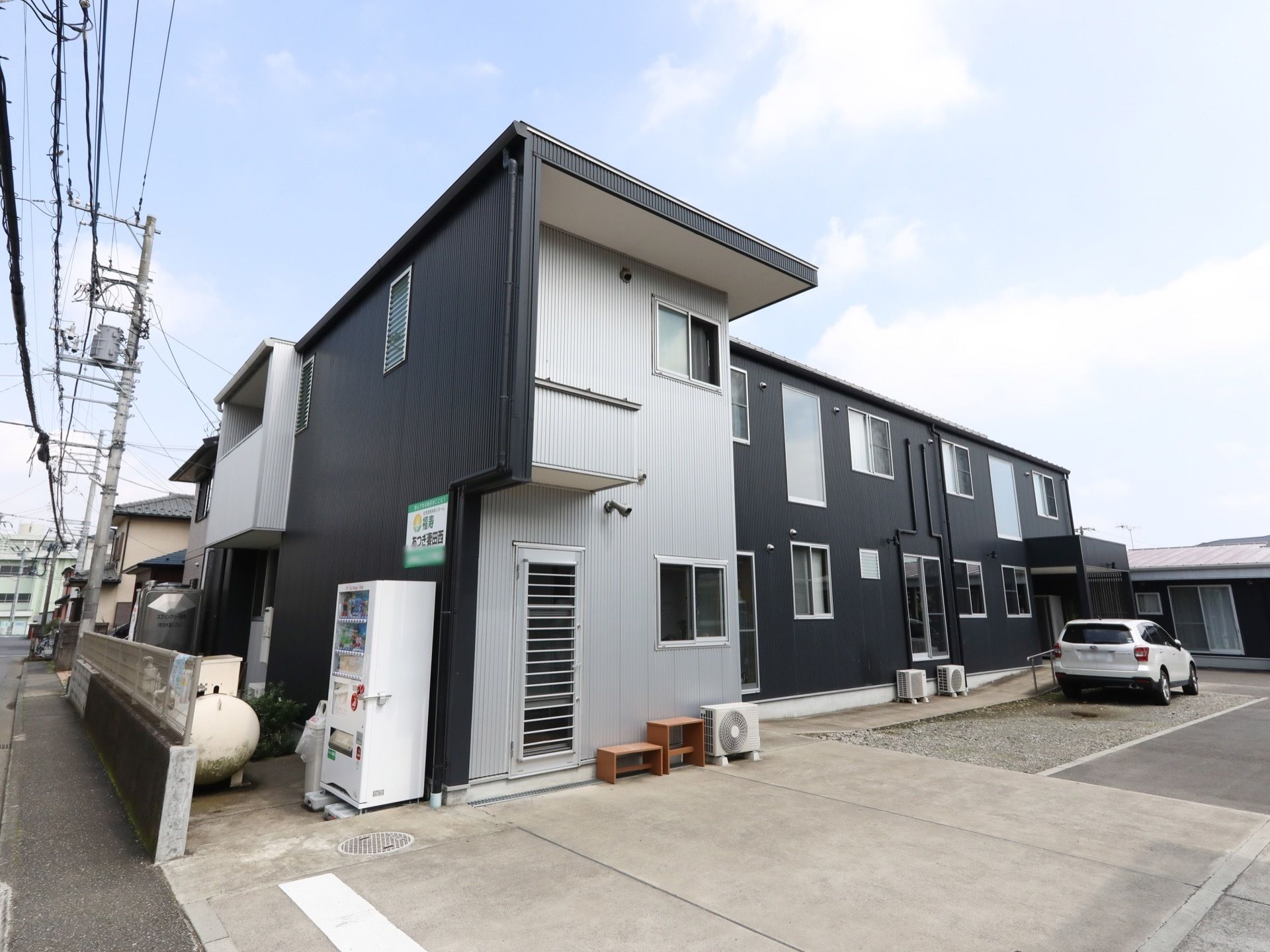 モノトーンを基調としたデザインの施設外観。道路に面した黒い壁には看板があり、その下には自動販売機が設置されている。