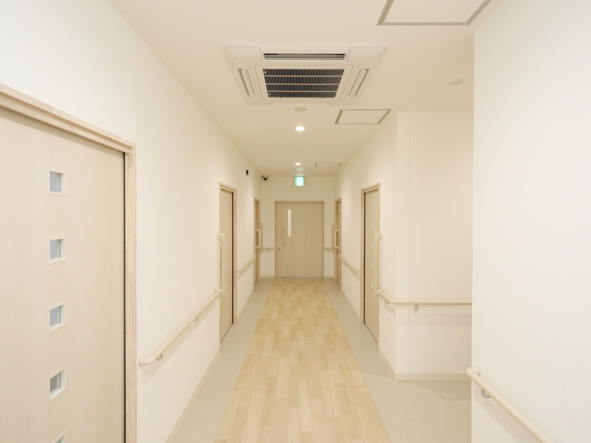 壁の両サイドに手すりが取り付けられている廊下で、床はフローリングとなっている。天井には照明と空調設備がある。