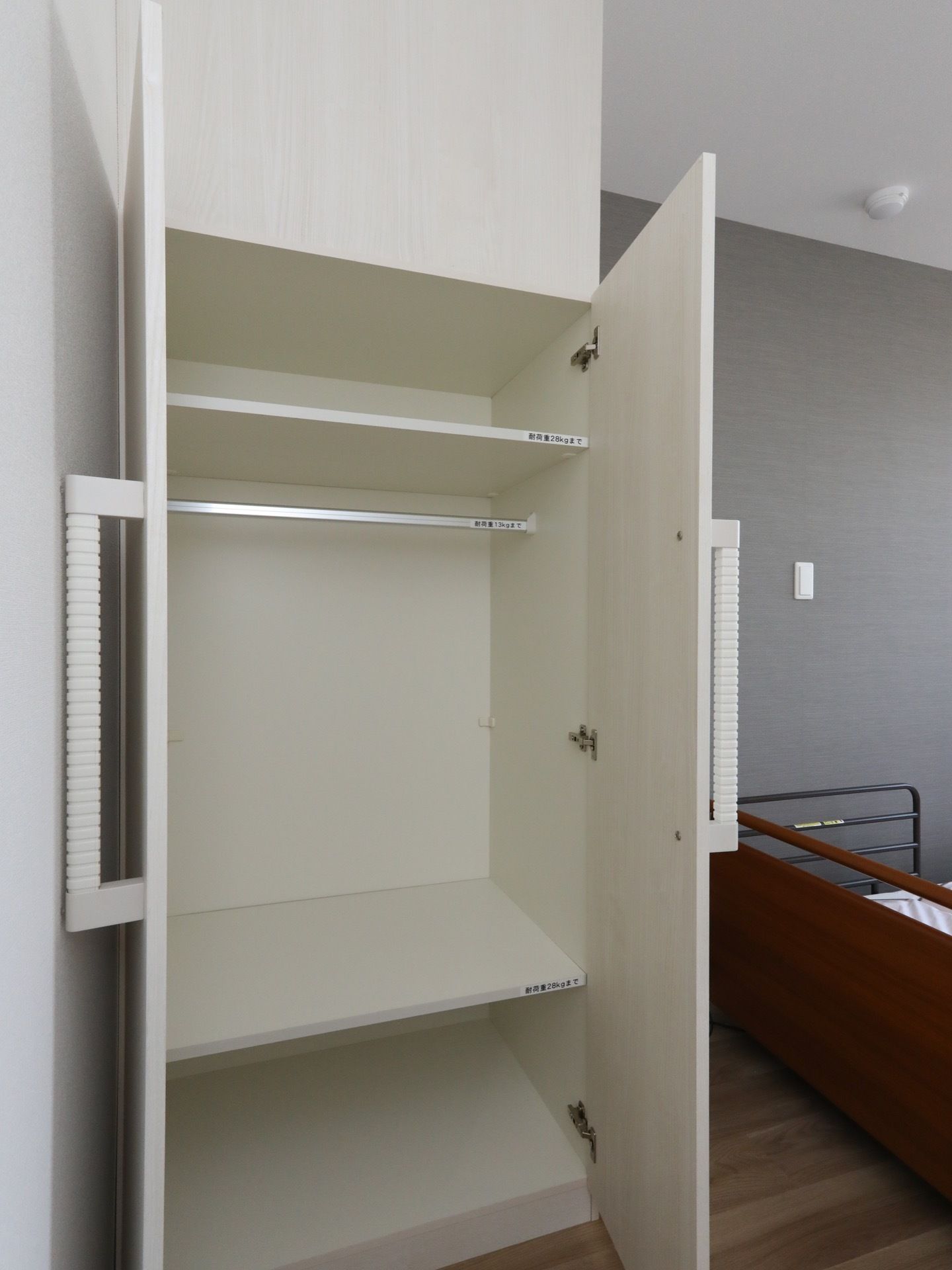 居室に備え付けられている両開きのクローゼットで、棚やポールがある。横にはベッドがある。