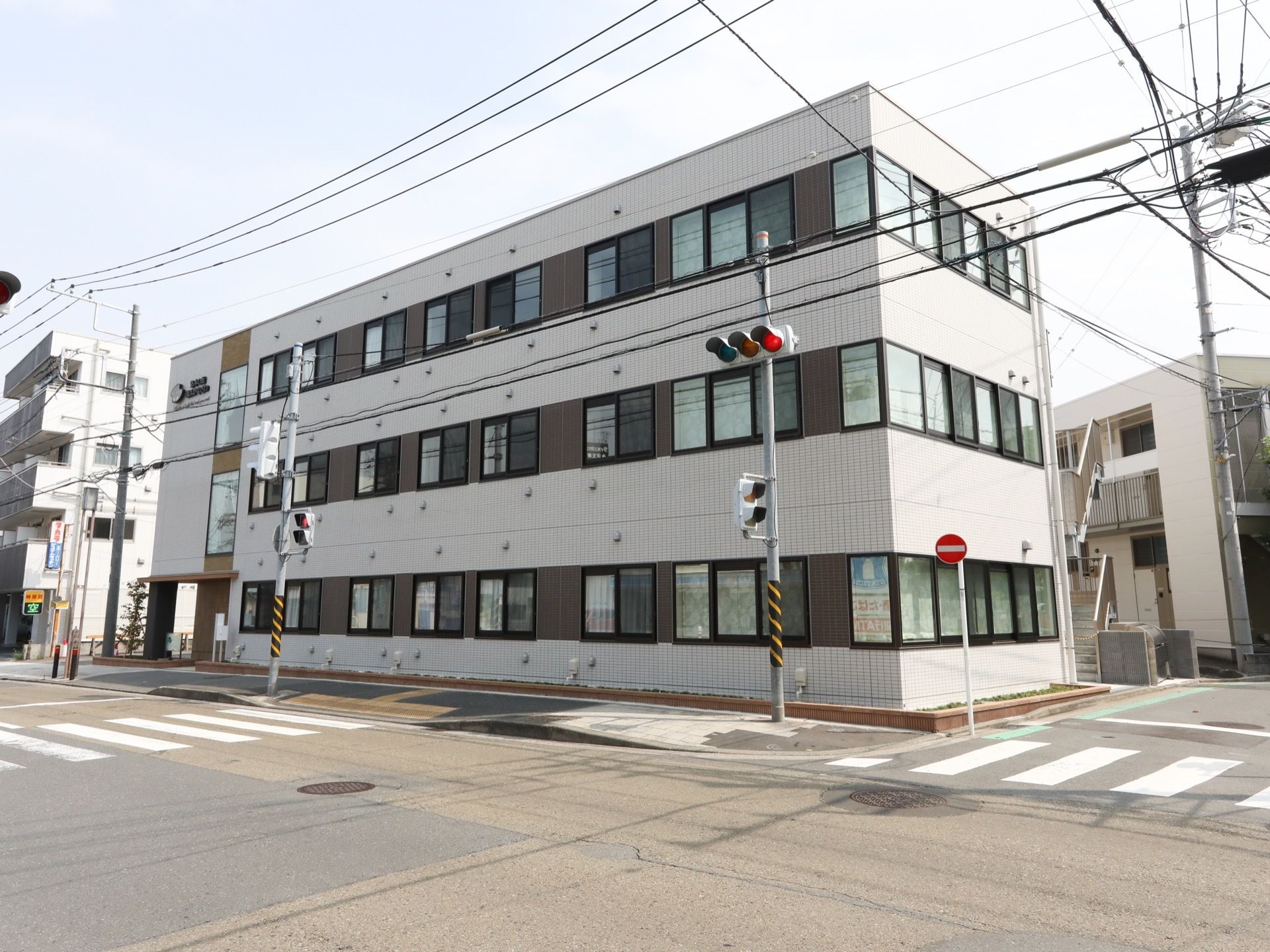 スタイリッシュな外観の四角い形のグループホームで、外壁は落ち着いた色合いとなっている。施設目の前には横断歩道がある。