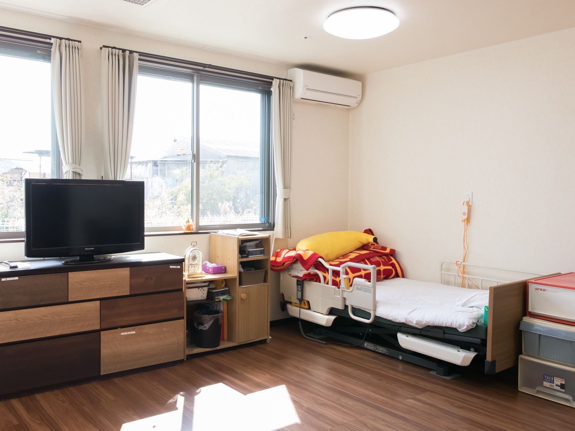 窓の手前に３段の棚があり、その上にテレビが設置されている。テレビの右側には置き時計やティッシュの箱などが置かれている。