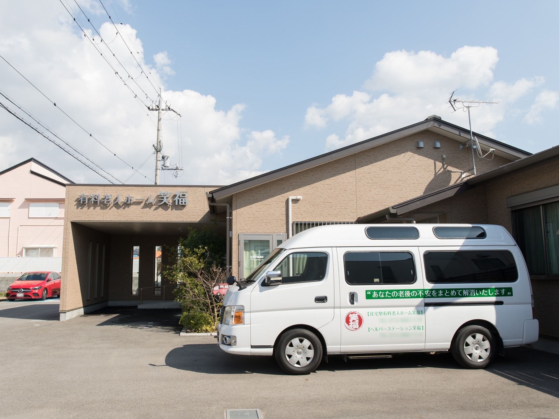 ベージュの外壁の１階建ての施設である。建物の前に白い車が１台停まっており、側面に施設名などが書かれている。