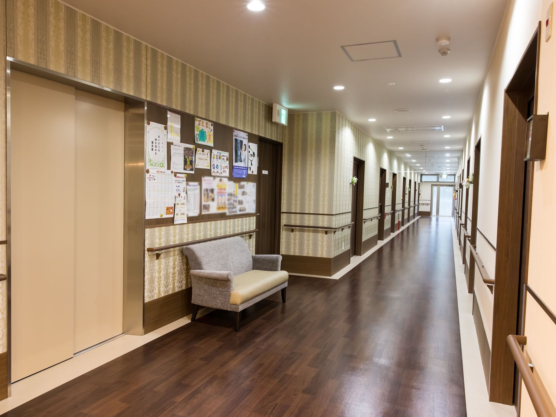 廊下の片側の壁が、縦のストライプ模様になっていて、天井に多くのダウンライトが設置されている。エレベーター乗降口の近くにソファが置かれている。