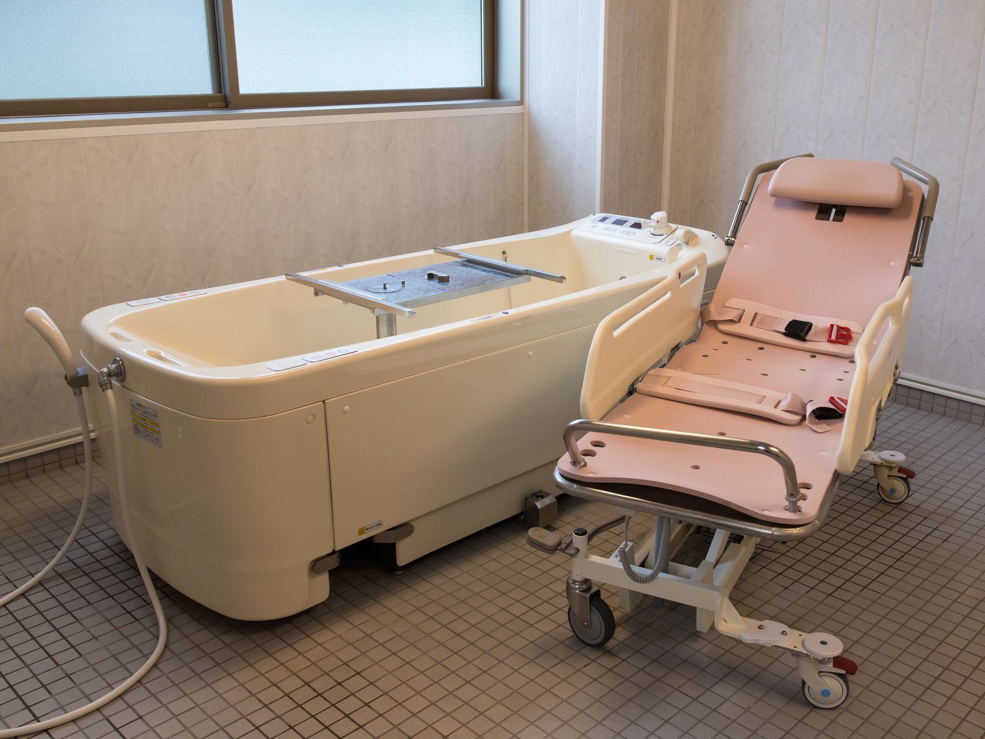 可動式のピンク色のストレッチャーが介護浴槽の隣に置かれている。浴槽内部でストレッチャーの受け具がせり上がっている。