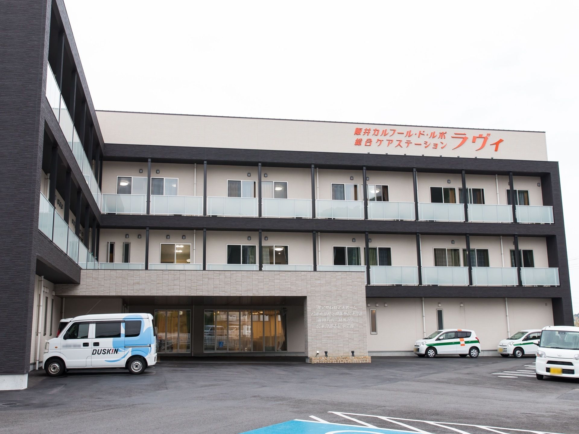 黒色の枠組みが特徴的な３階建ての施設である。外壁の一番上の部分にオレンジ色の文字で施設名称が表示されている。