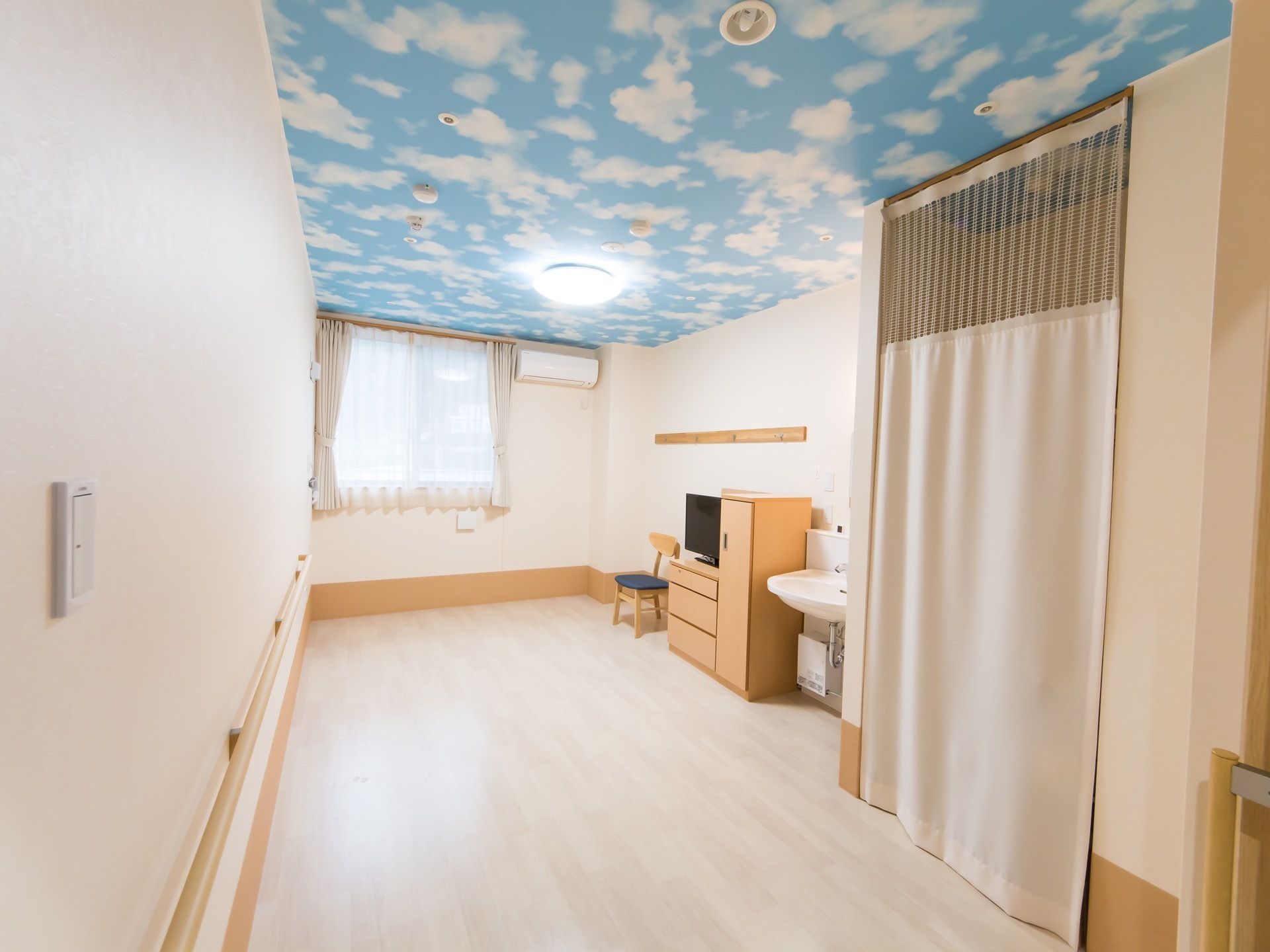 居室の天井に、青空に白い雲が浮かんでいる絵が描かれている。腰高の窓があり、その右側の壁の上の方にエアコンを設置。