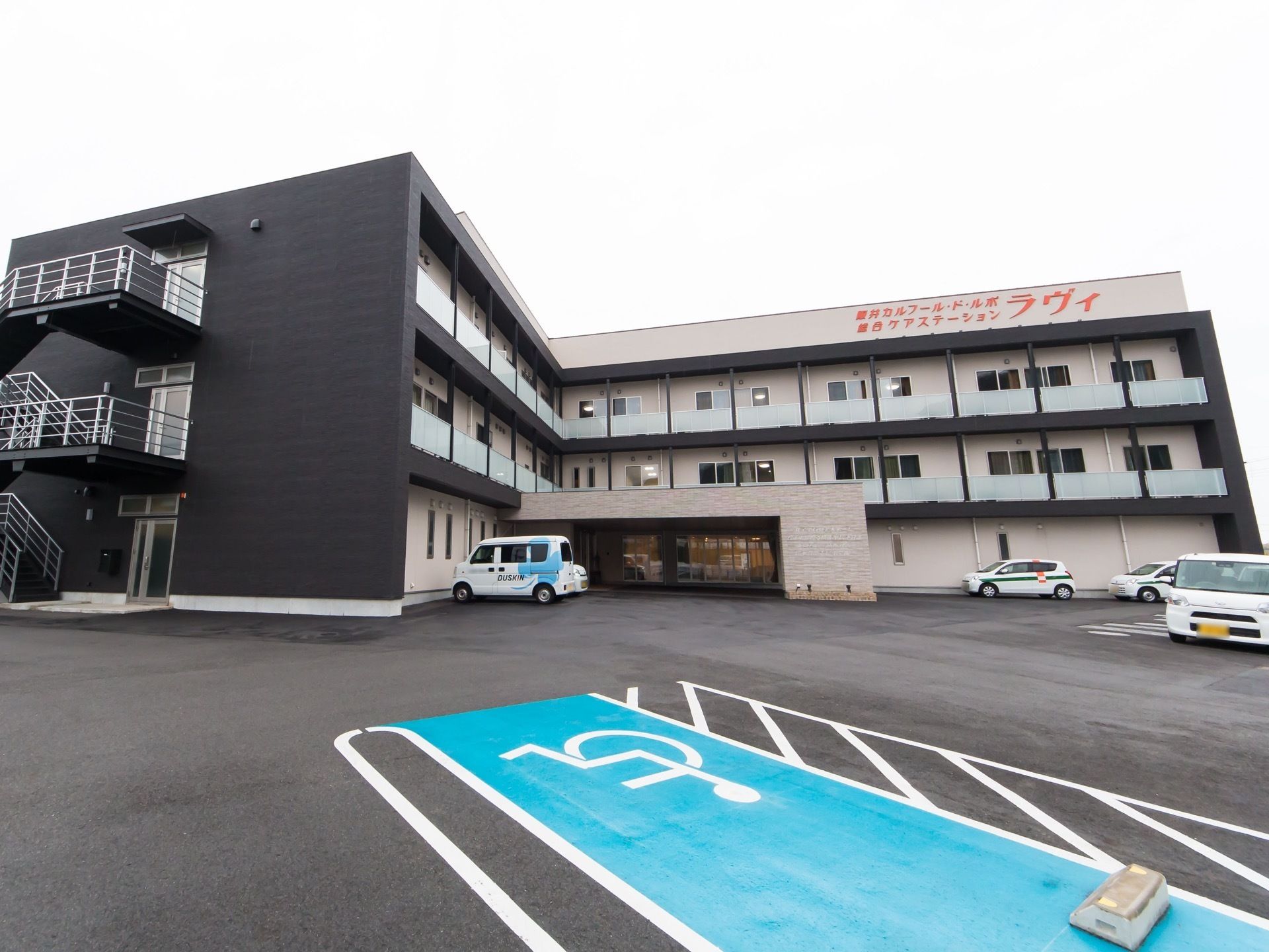 白色と黒色を基調とした外壁を持つ３階建ての施設である。車椅子のマークが描かれた駐車スペースが水色で塗られている。