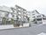 4階建ての建物は全体が白の基調となっている。また、手前には駐車場や駐輪場に停まっている車や自転車も見える。