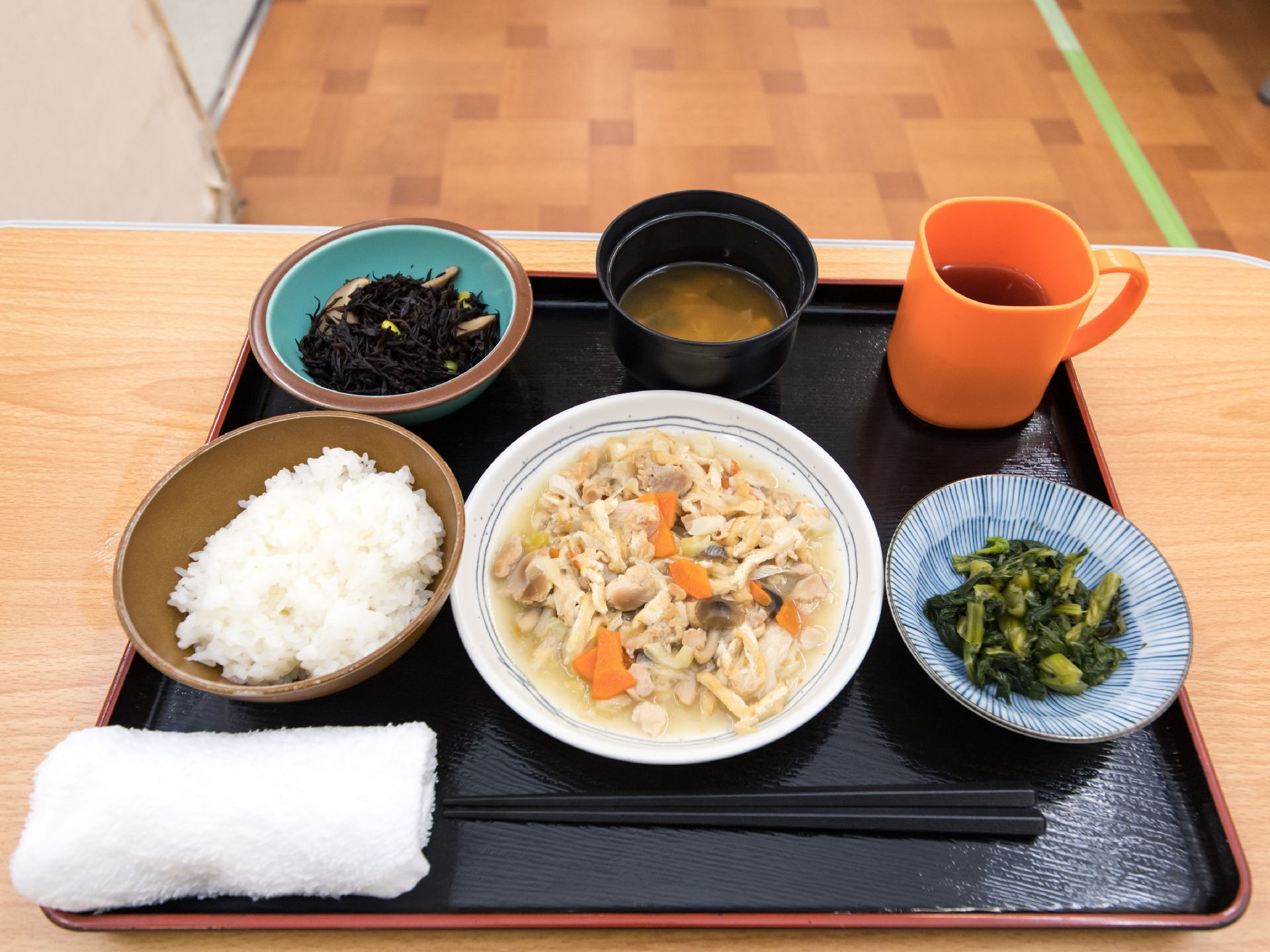 栄養バランスの良い食事と飲み物が黒いトレーにのせられている。白米とお味噌汁を中心に煮物や和え物が並べられている。