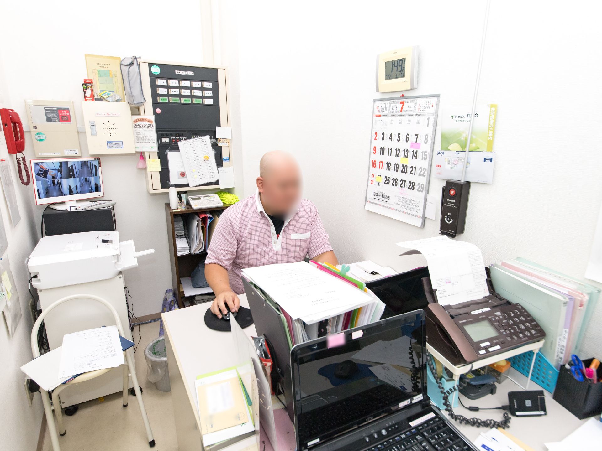 事務室には、コピー機やノートパソコン、ファックス付き電話機などの情報機器が設置されている。男性のスタッフがデスクで作業をしている。