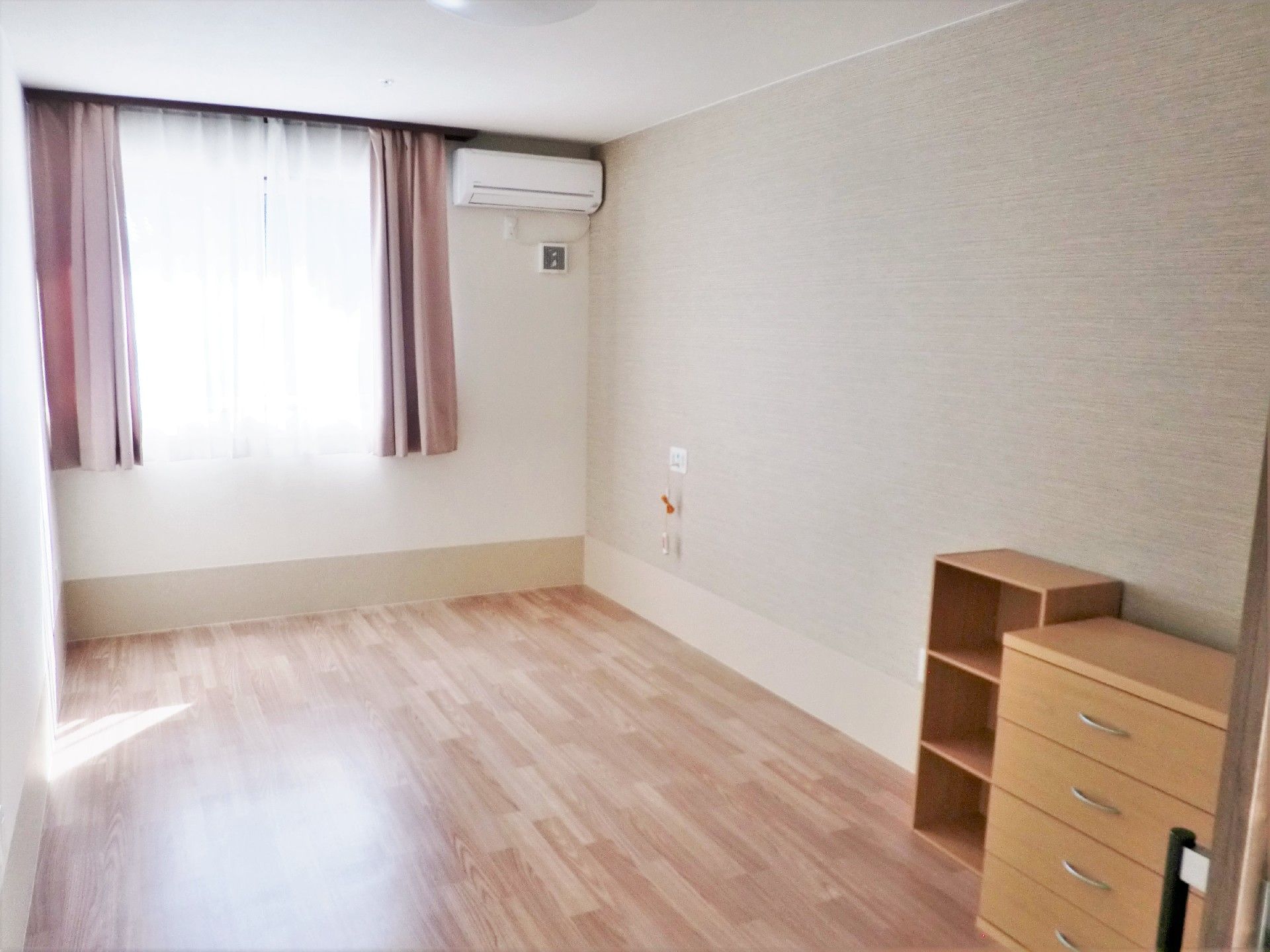 フローリングの小さな部屋の全景。奥のほうに窓とエアコン、手前にカラーボックスとたんすが置いてある部屋
