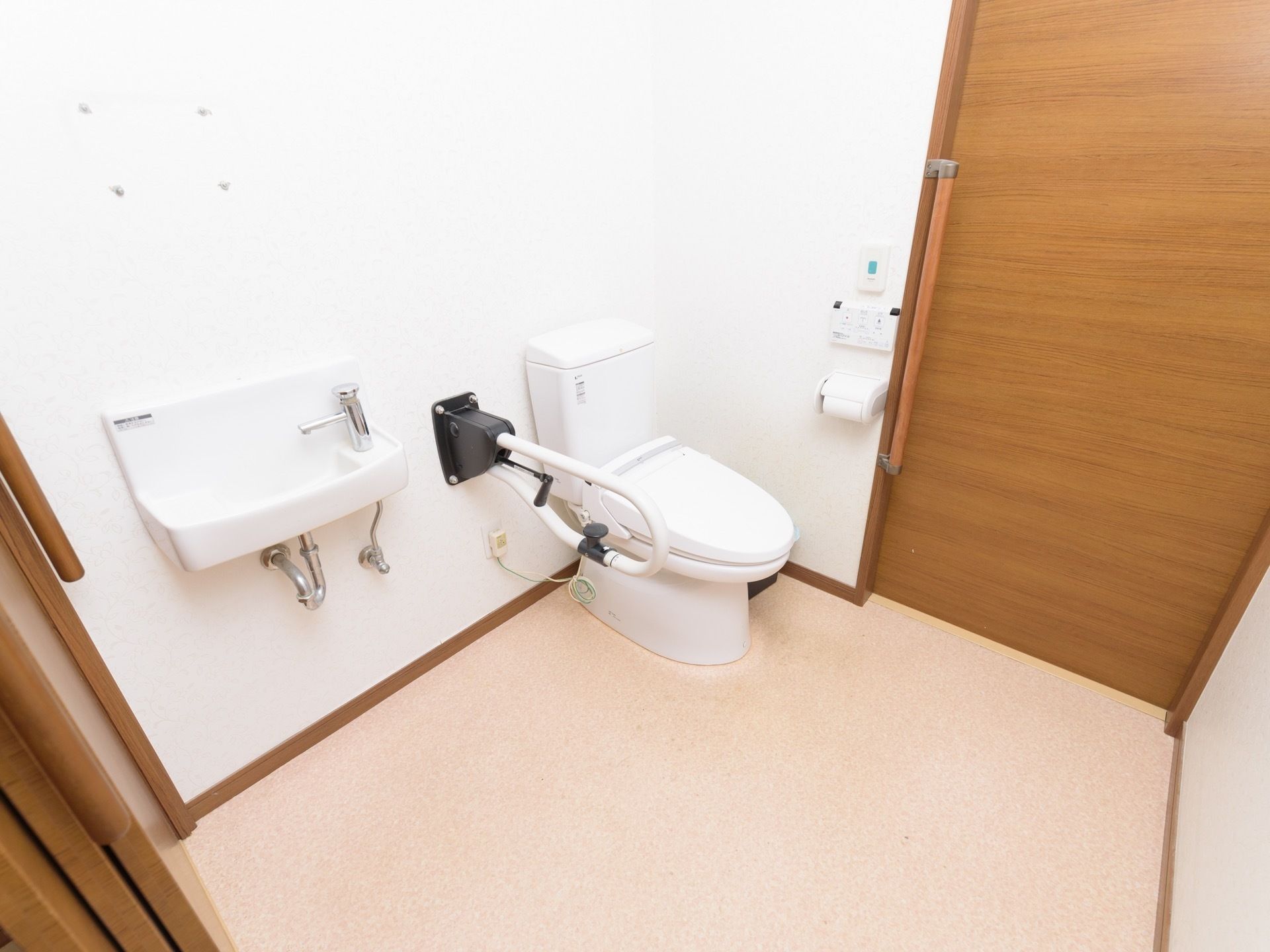 広いスペースにトイレが設置されている。ふたつきの便座の横には、可動式の手すりがある。壁設置式の洗面台がついている。