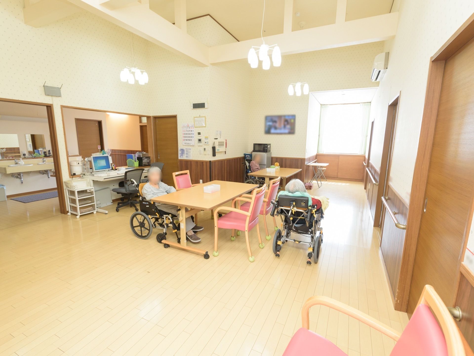 休憩スペースがある。テーブルが２つと、椅子がいくつか置かれている。車椅子をご利用の入居様が３名集まっている。