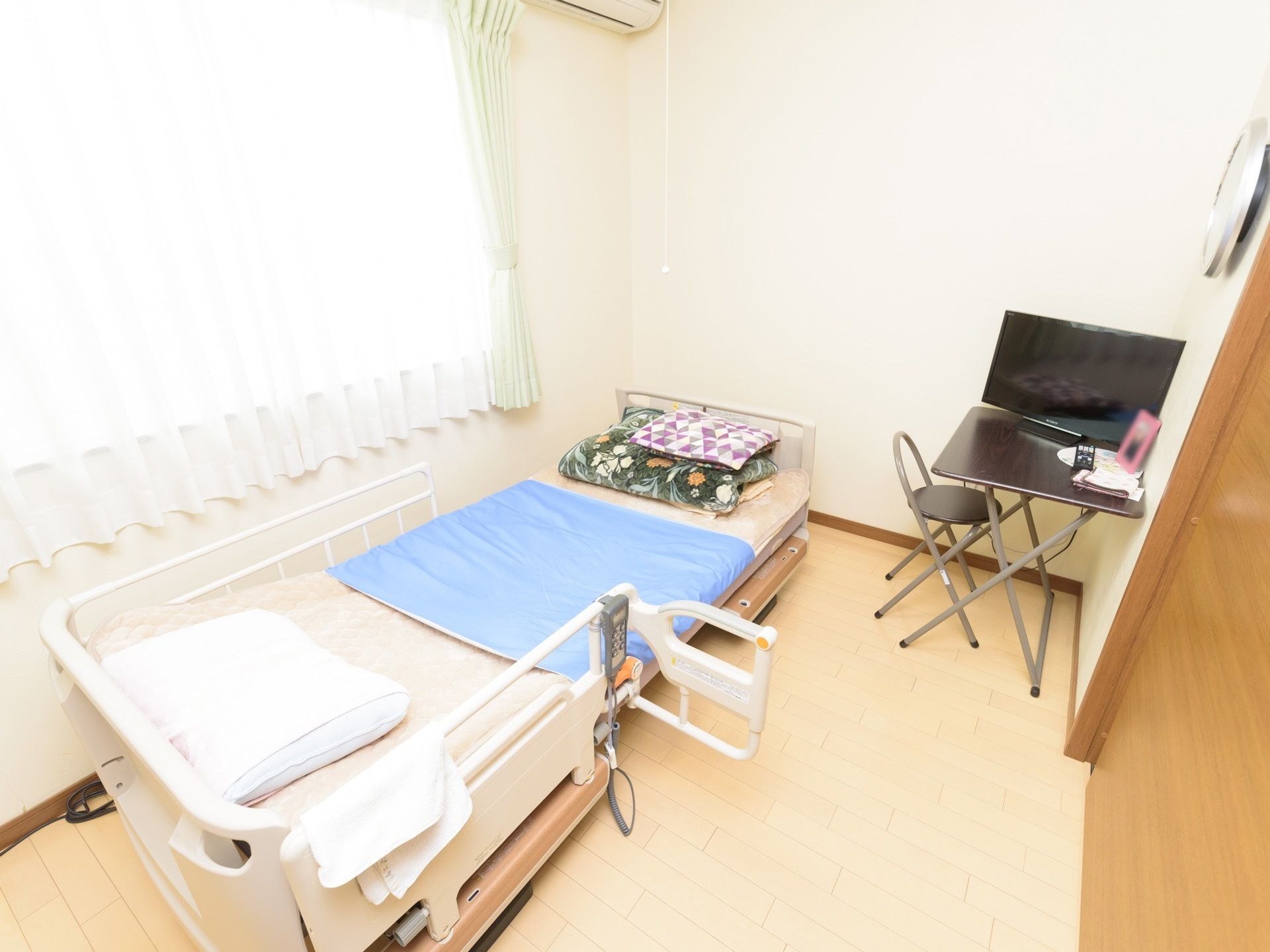 居室には大きな窓があり、レースのカーテンが閉められている。転落防止柵月のベッドがあり、小さな台の上にテレビが置かれている。