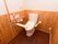 余裕ある広さのトイレの個室。周囲の壁は木目になっていて、温水洗浄便座と手すりなどを備え付けた洋式トイレが１つ設置されている。