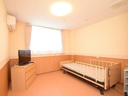ベッドやテレビ、たんすにエアコンなどがそろった居室。窓には白いカーテンが付いている。