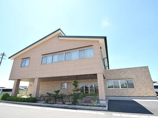 ベージュ系の2階建ての建物。駐車場が付き、玄関周りまで2階部分があるため、屋根の役割をしている。