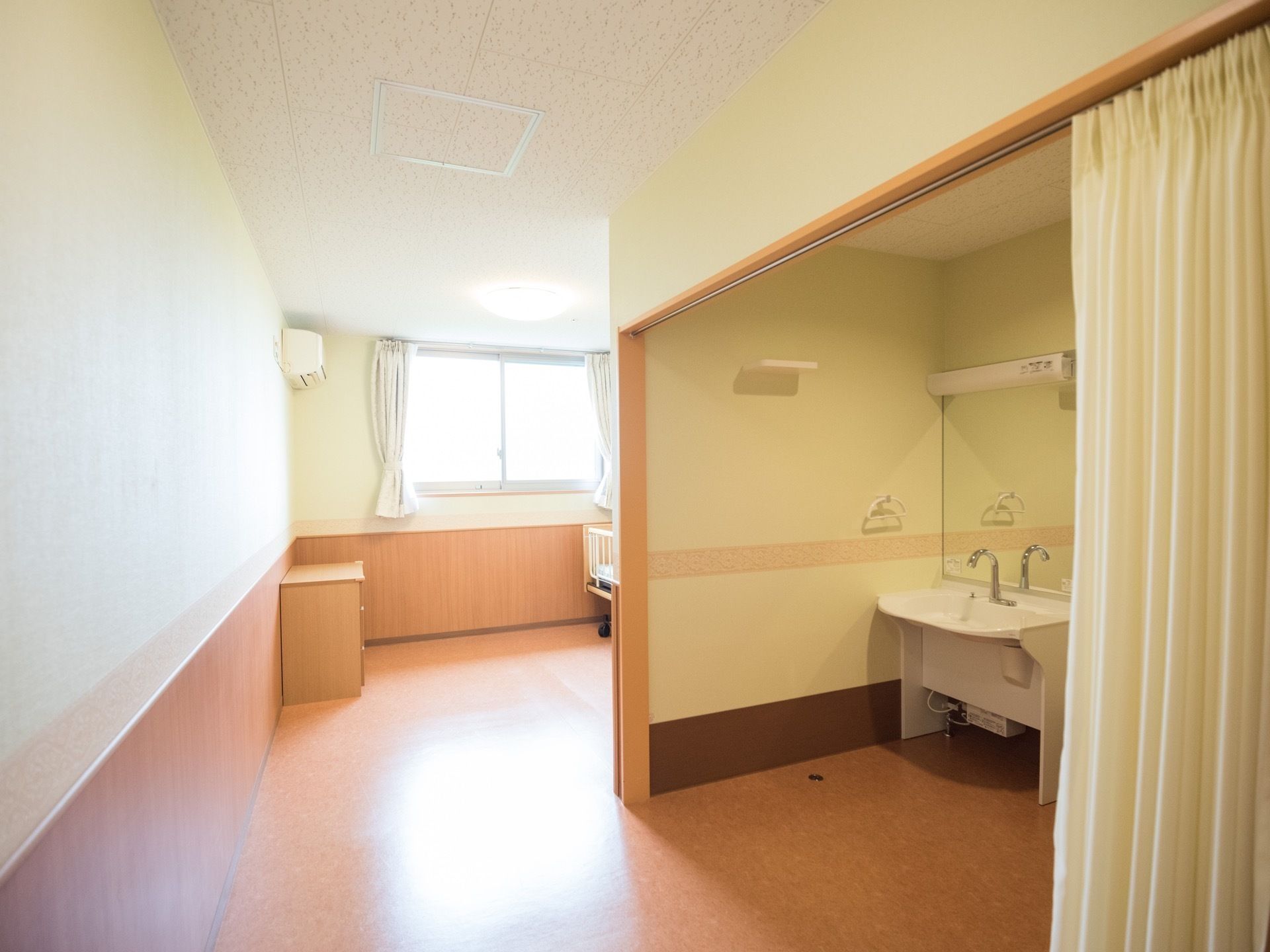 居室に入ると右手に洗面台とトイレがある。奥にはエアコンの付いた洋室があり、介護用ベッドが一台設置されている。