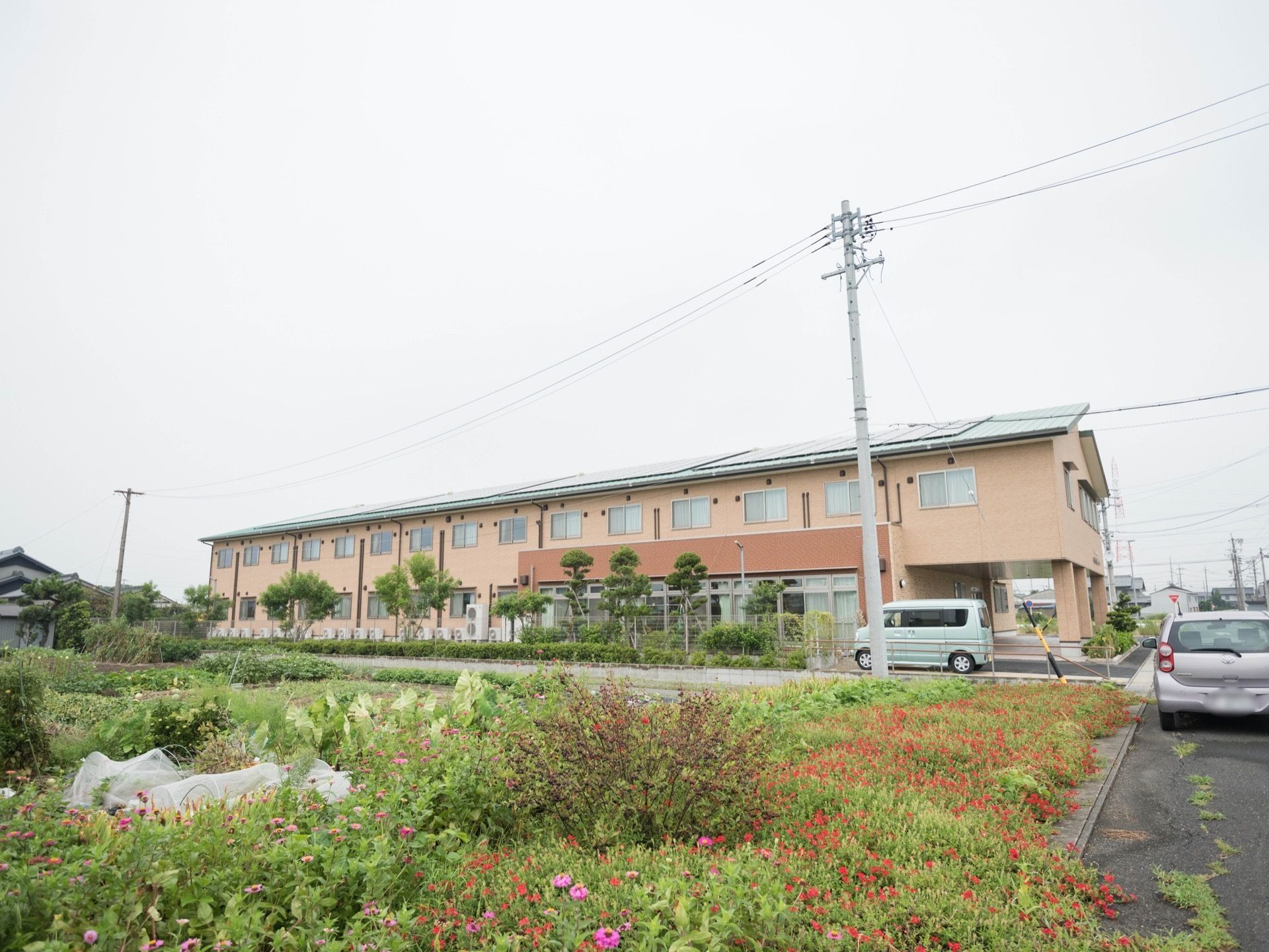 植栽に囲まれた敷地に、薄いオレンジ色の外壁をした2階建ての住宅が建っている。周辺は畑や戸建ての多い住宅地になっている。