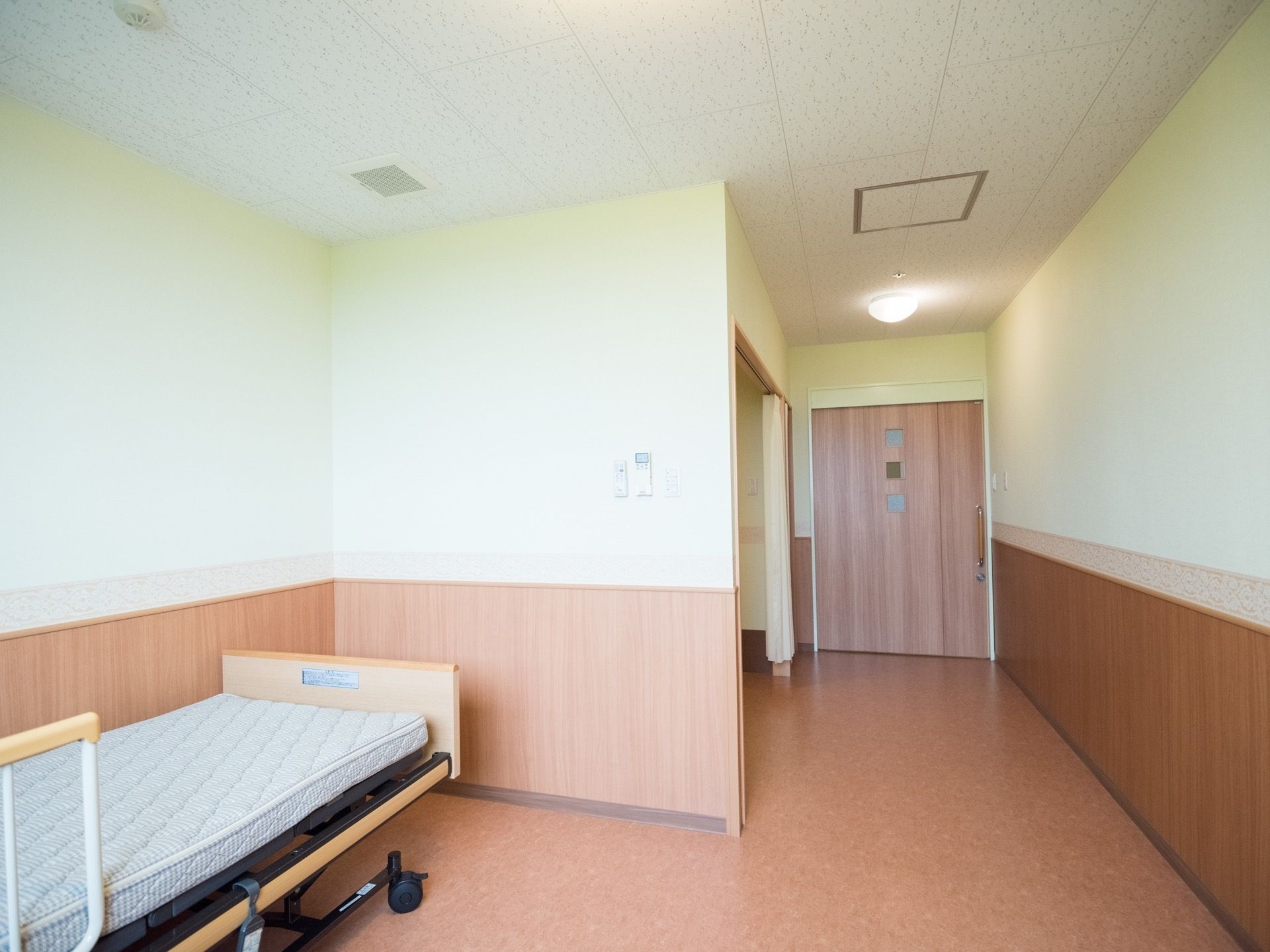 電動ベッドが一台置かれた居室がある。入り口は木目調の引き戸であり、壁も白や木目調の柔らかいデザインで統一されている。