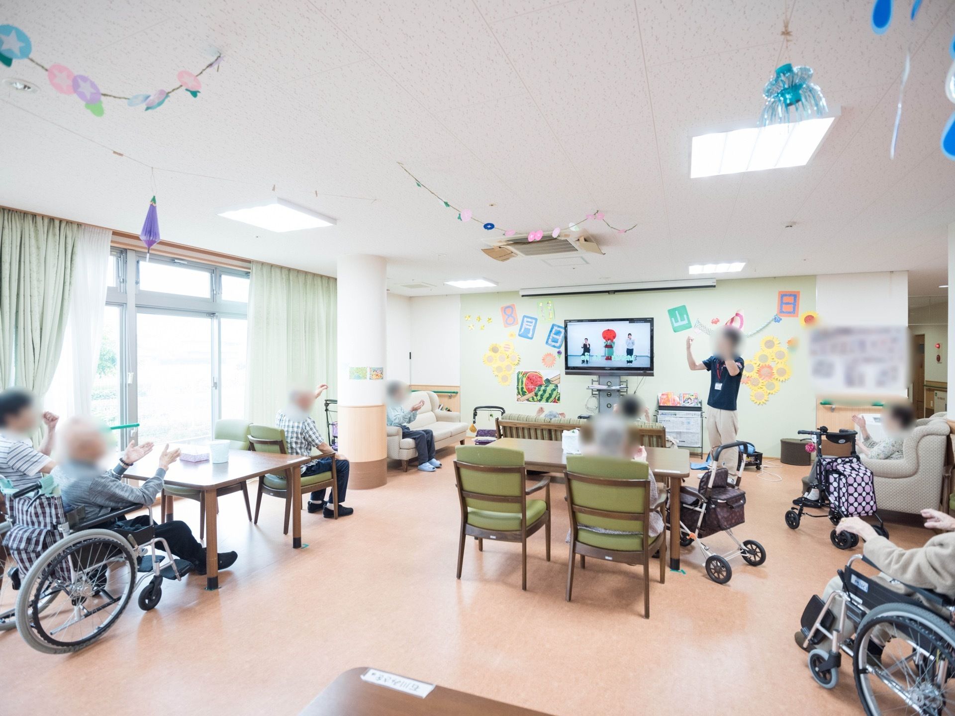 空調の付いた広いフロアで、入居者様が集まっている。スタッフと一緒にテレビ画面を観ながら体操を行っている。