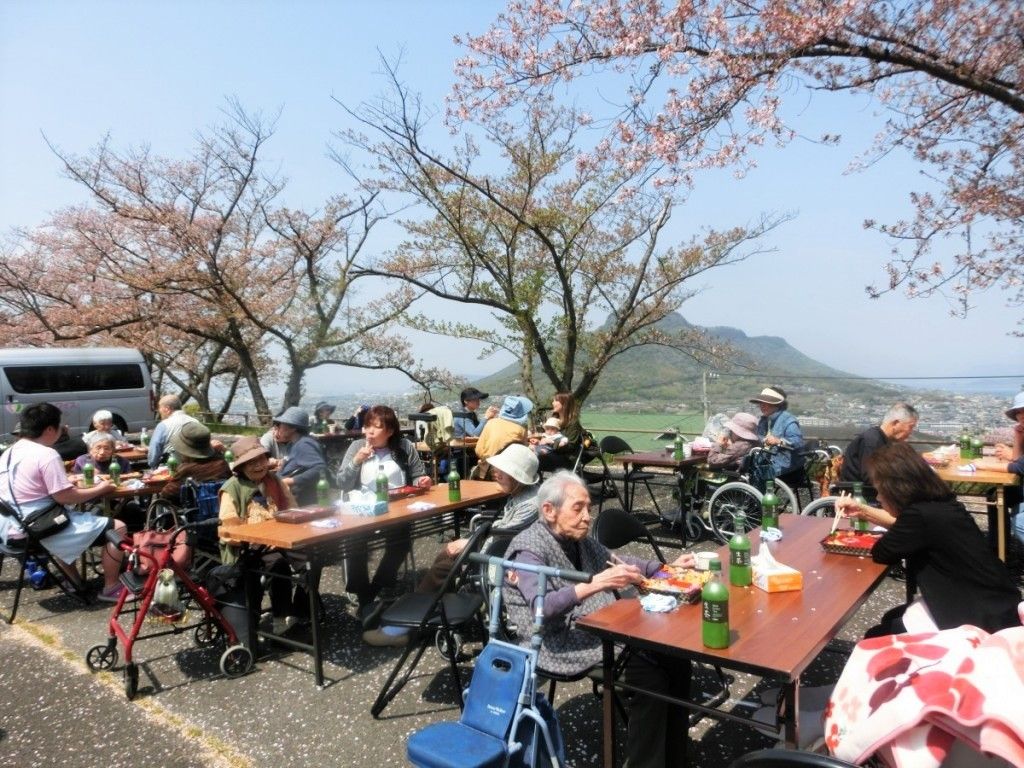 桜の木の下で花見を楽しむ入居者とスタッフ。大きなテーブルが複数並べられている。