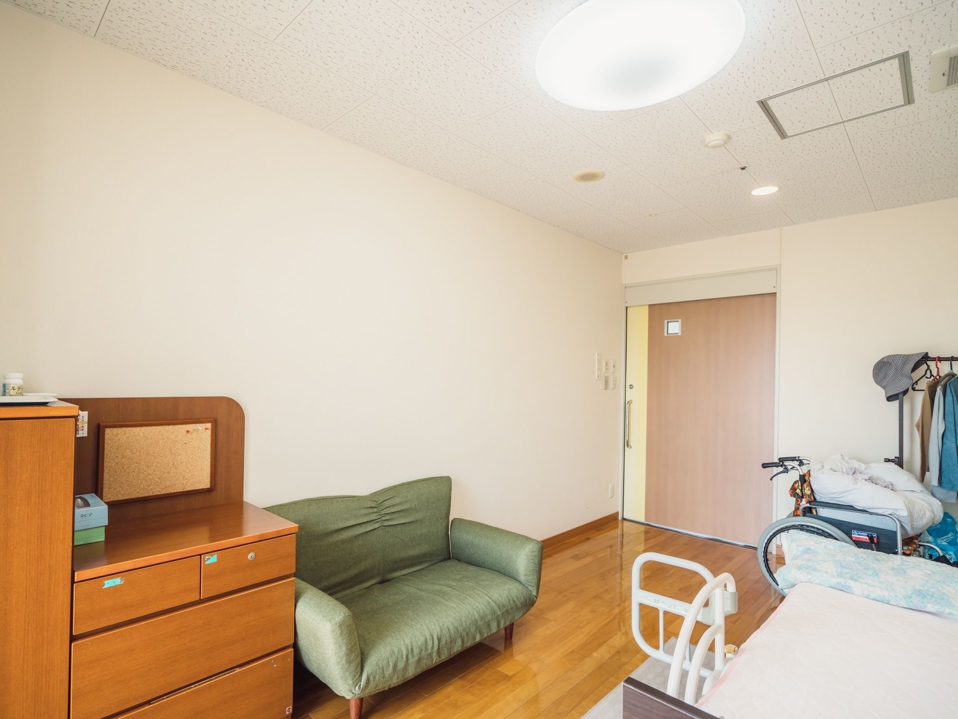 左側には木製の収納棚と緑色のソファが設置されている。右側にはベッドがあり、車椅子と衣類が掛けられたハンガーラックが置いてある。