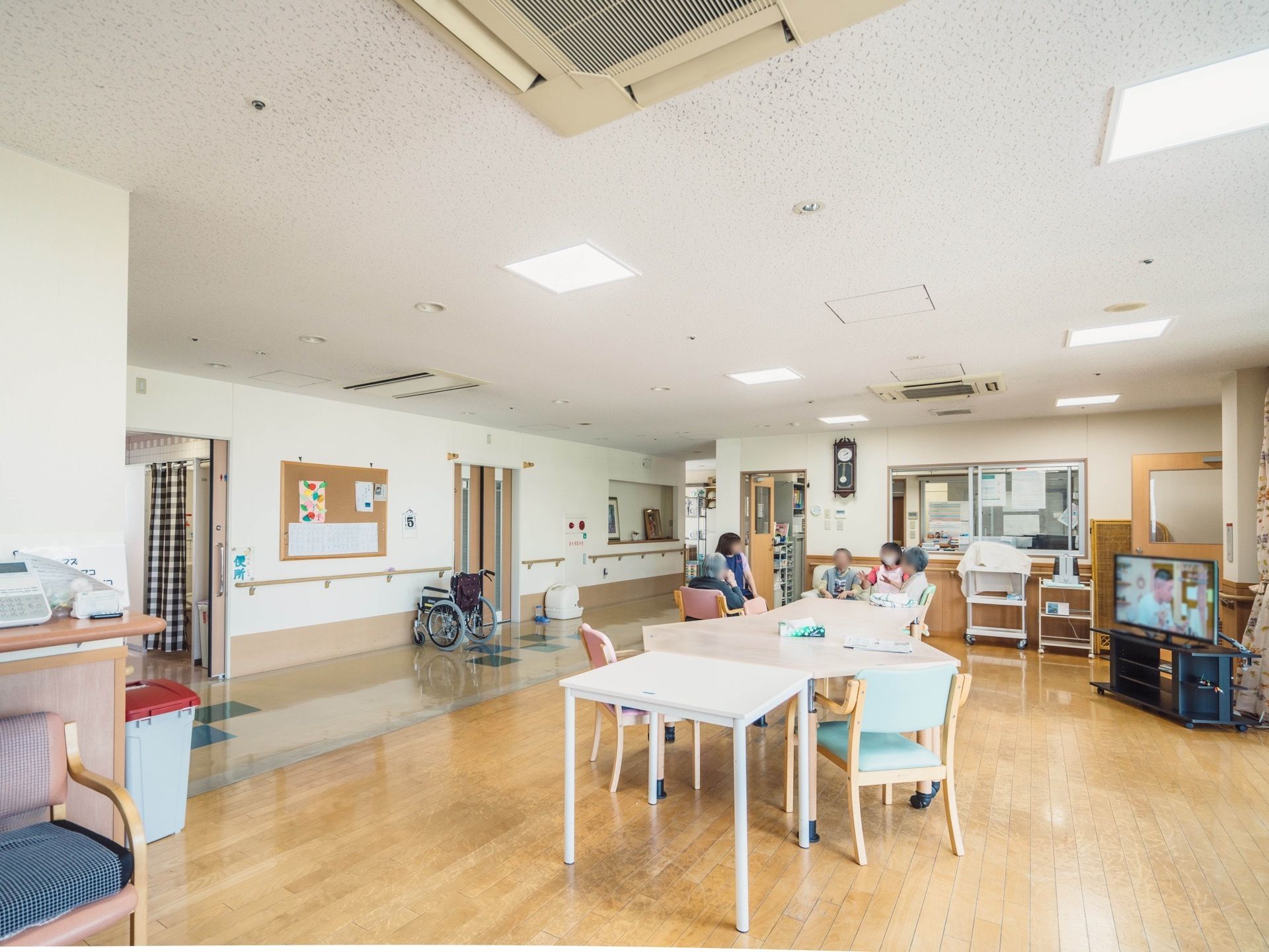 テーブルや椅子が配置された広間で、液晶テレビが稼働している。奥の方で複数の人が向かい合って話している様子が見える。