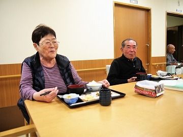 ダイニングテーブルについて食事を楽しんでいる男女の入居者。それぞれの前には黒いトレーに乗ったご飯やおかずがある。