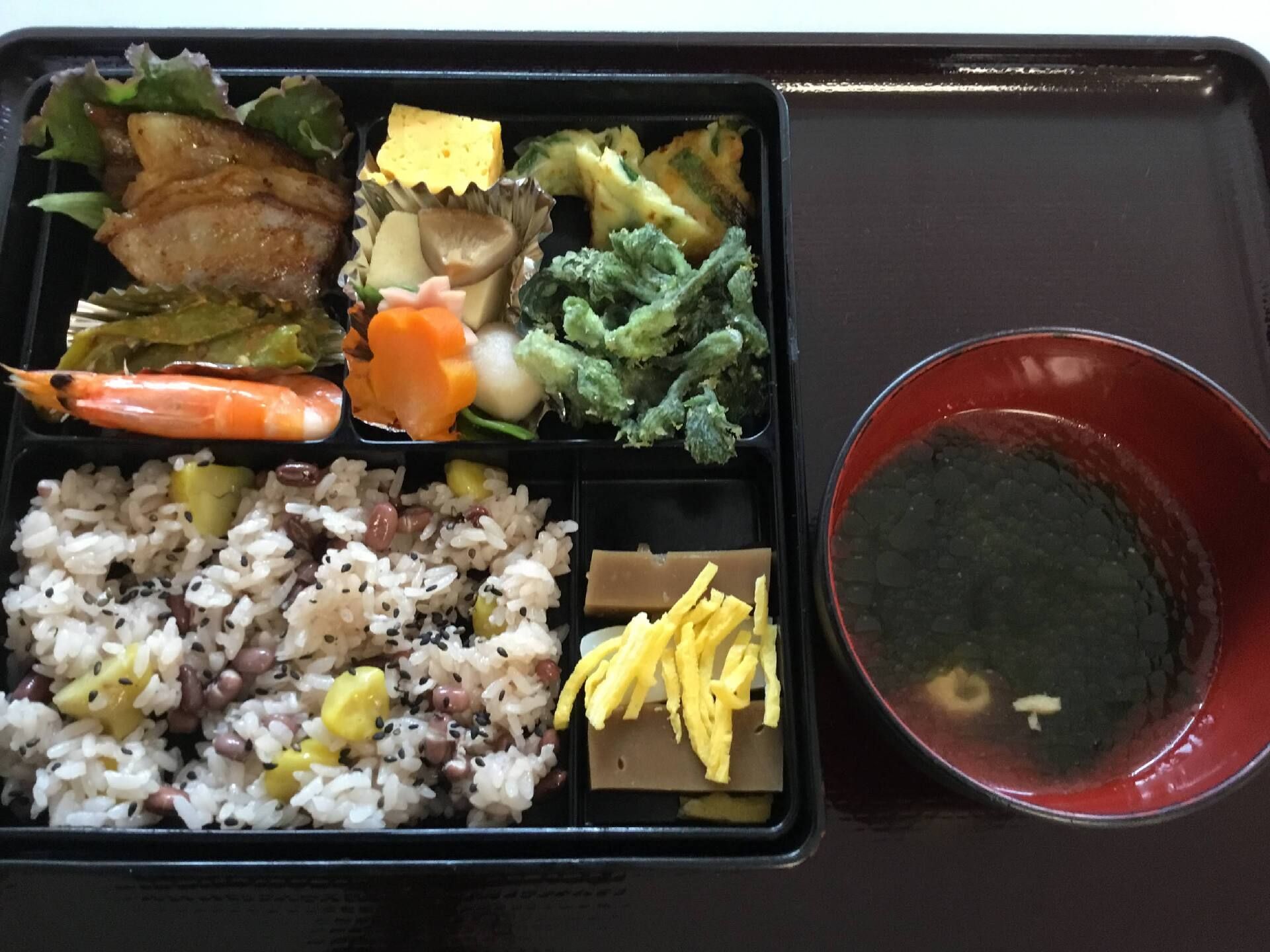 サツマイモの炊き込みご飯には黒ゴマが振りかけられ、魚や煮物、野菜の和え物などが詰められた弁当風の食事。汁物も添えられている。